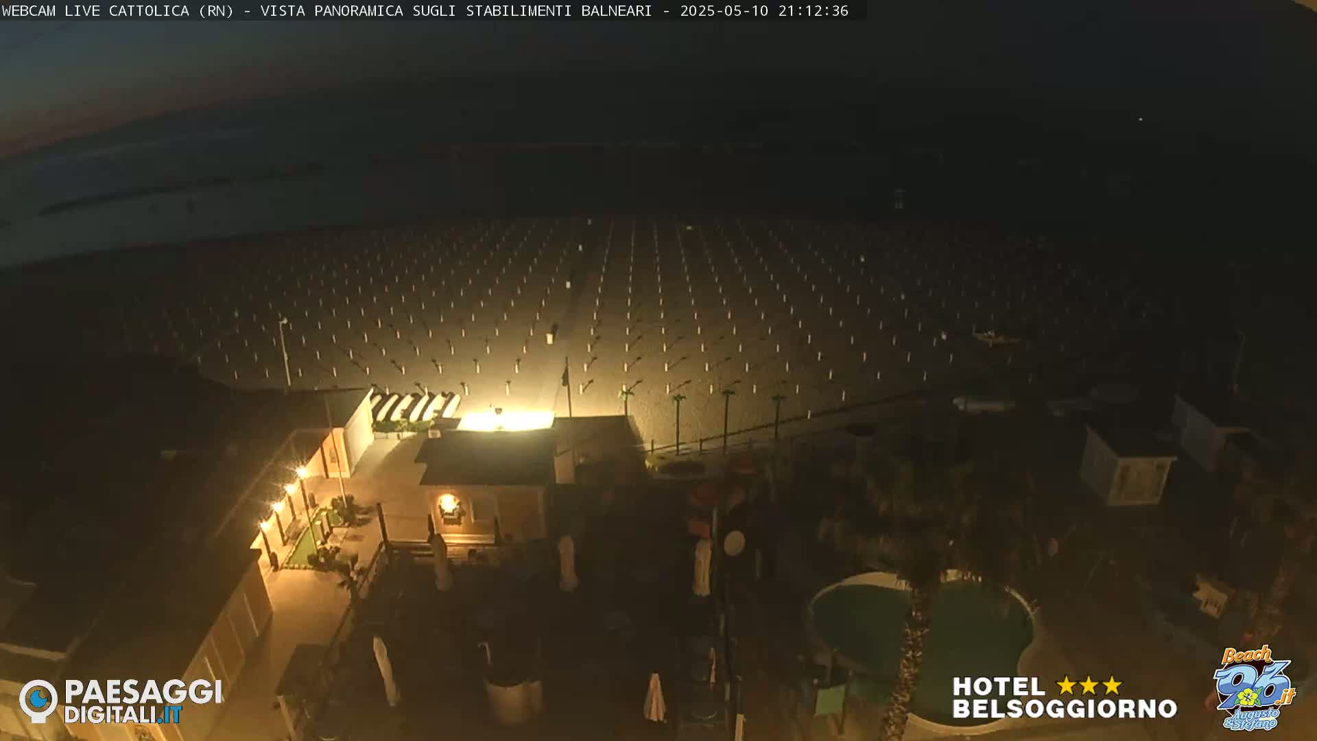 At night, a beach with rows of beach umbrellas is illuminated, next to a building with a pool.