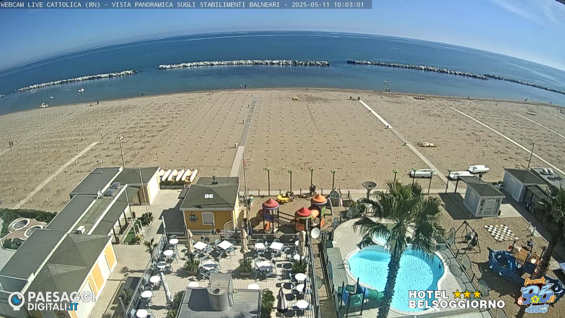 An aerial view shows a sandy beach with rows of umbrellas, beach equipment, a small playground, a swimming pool, and outdoor seating at a beachfront establishment on a sunny day.