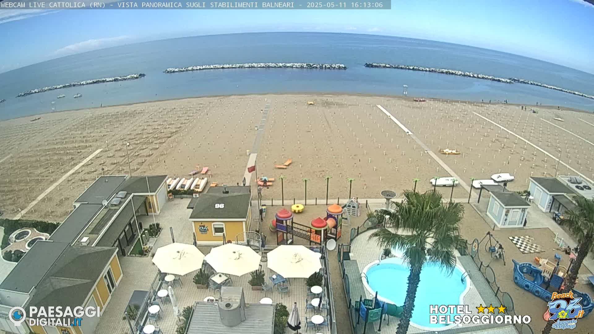 A mostly sunny day at a beach, showing a sandy beach with rows of umbrellas, beachside buildings, a swimming pool, and playground equipment.