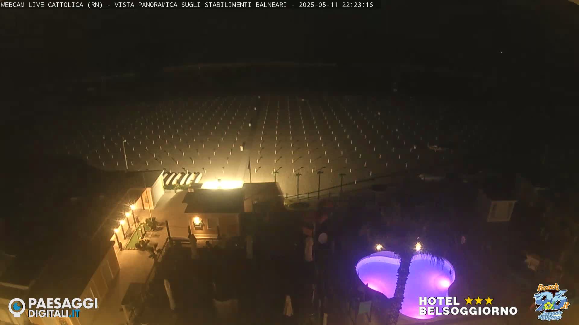 A nighttime, aerial view shows a dark beach with numerous evenly spaced lights, a lit building, and a purple-lit swimming pool.