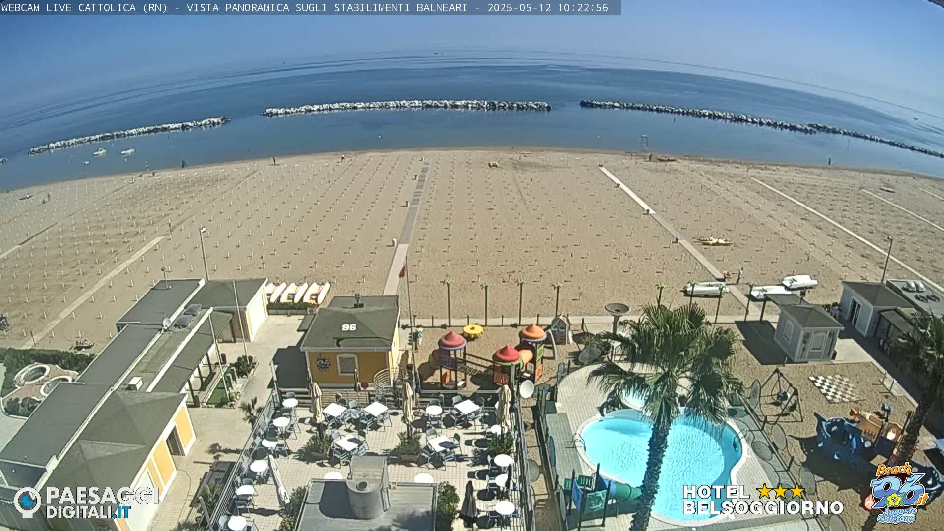 A beach with rows of umbrellas and beach chairs, a hotel with a pool and playground, under a sunny sky.