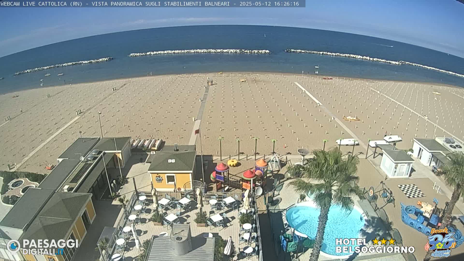 An overhead view of a beach with numerous umbrellas and lounge chairs, a playground, a swimming pool, and beachside buildings on a sunny day.