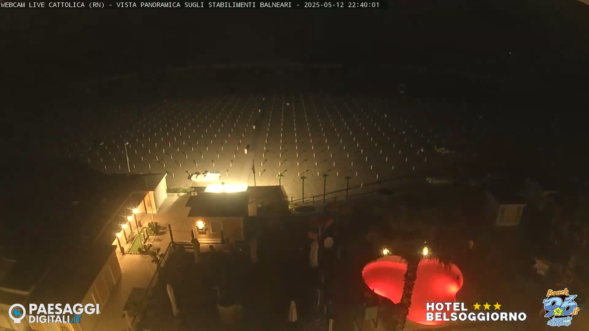 A nighttime aerial view shows a beach with rows of beach umbrellas and a building with a red-lit pool.