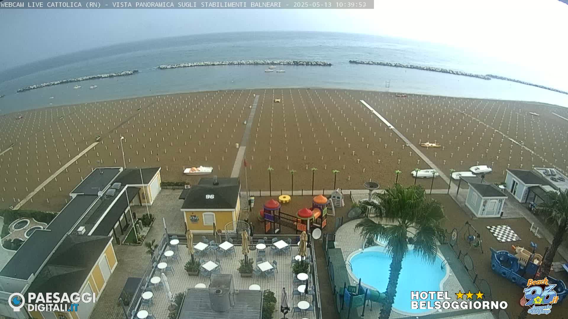 An overcast day reveals an empty sandy beach with rows of beach umbrellas, a small swimming pool, and a playground at a beachfront resort.