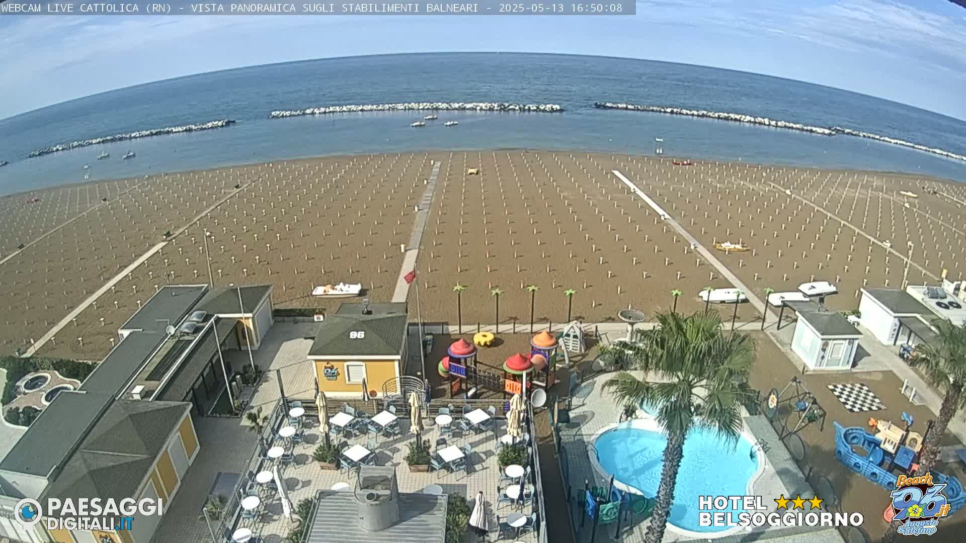 A sandy beach with rows of umbrellas and beach chairs is seen from an elevated vantage point, overlooking a calm ocean under a mostly clear sky;  a swimming pool, playground, and beachside restaurant are also visible.