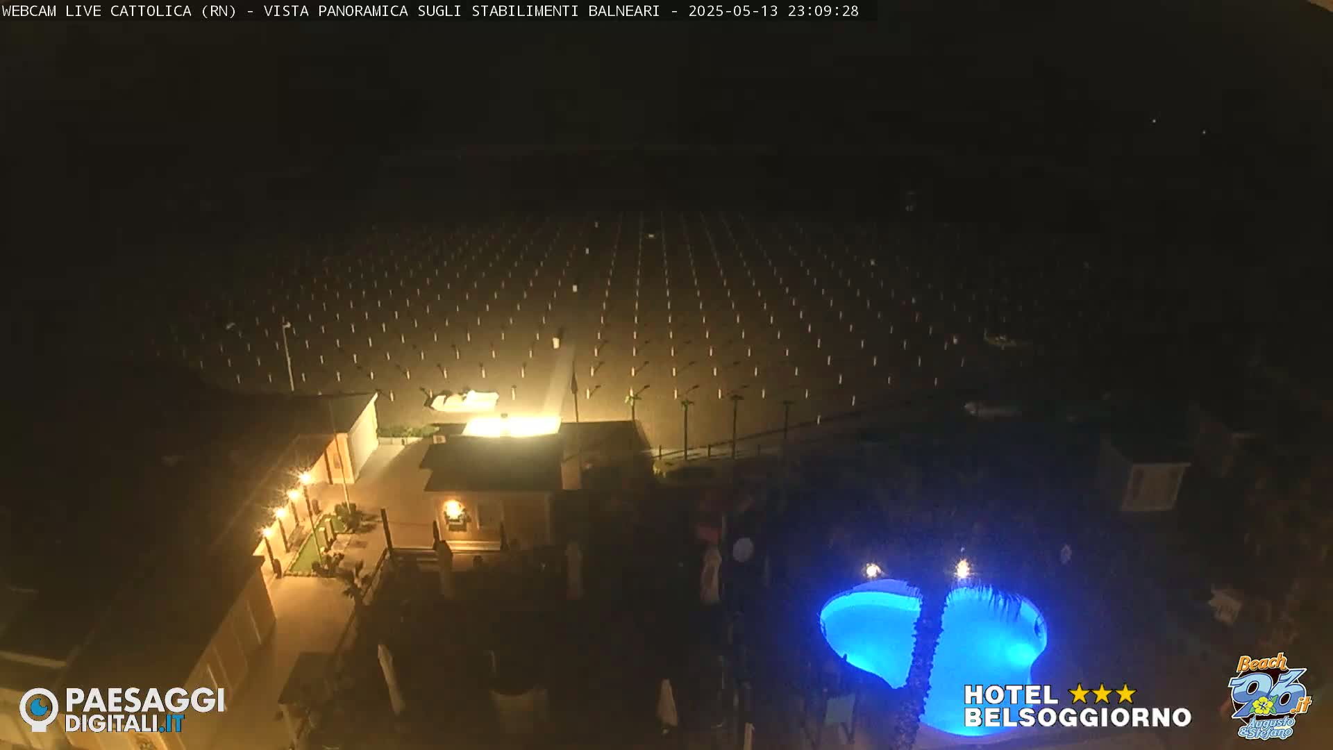 A nighttime aerial view shows a beach with numerous evenly spaced lights, a building with illuminated exterior, and a blue-lit swimming pool.