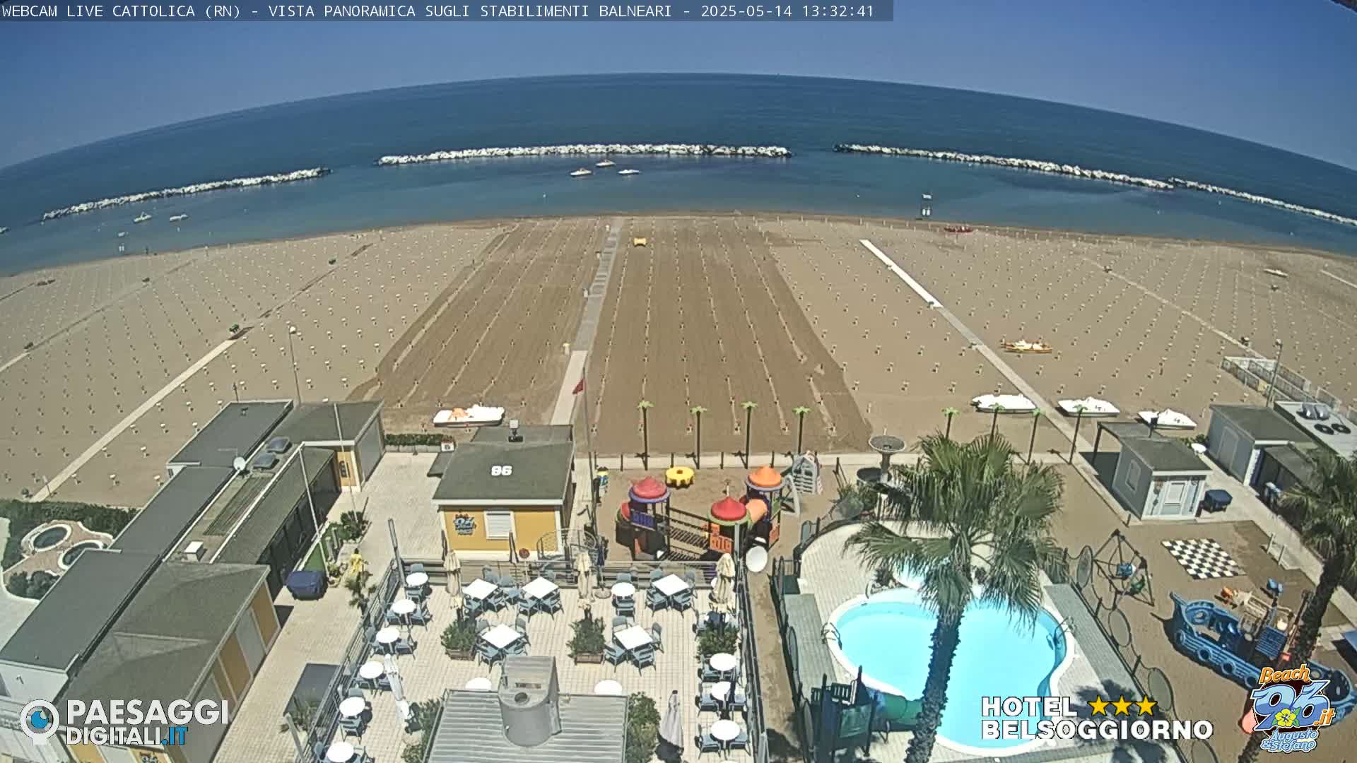 An aerial view shows a beach with rows of umbrellas and beach chairs, a swimming pool, a playground, and beachside buildings on a sunny day.