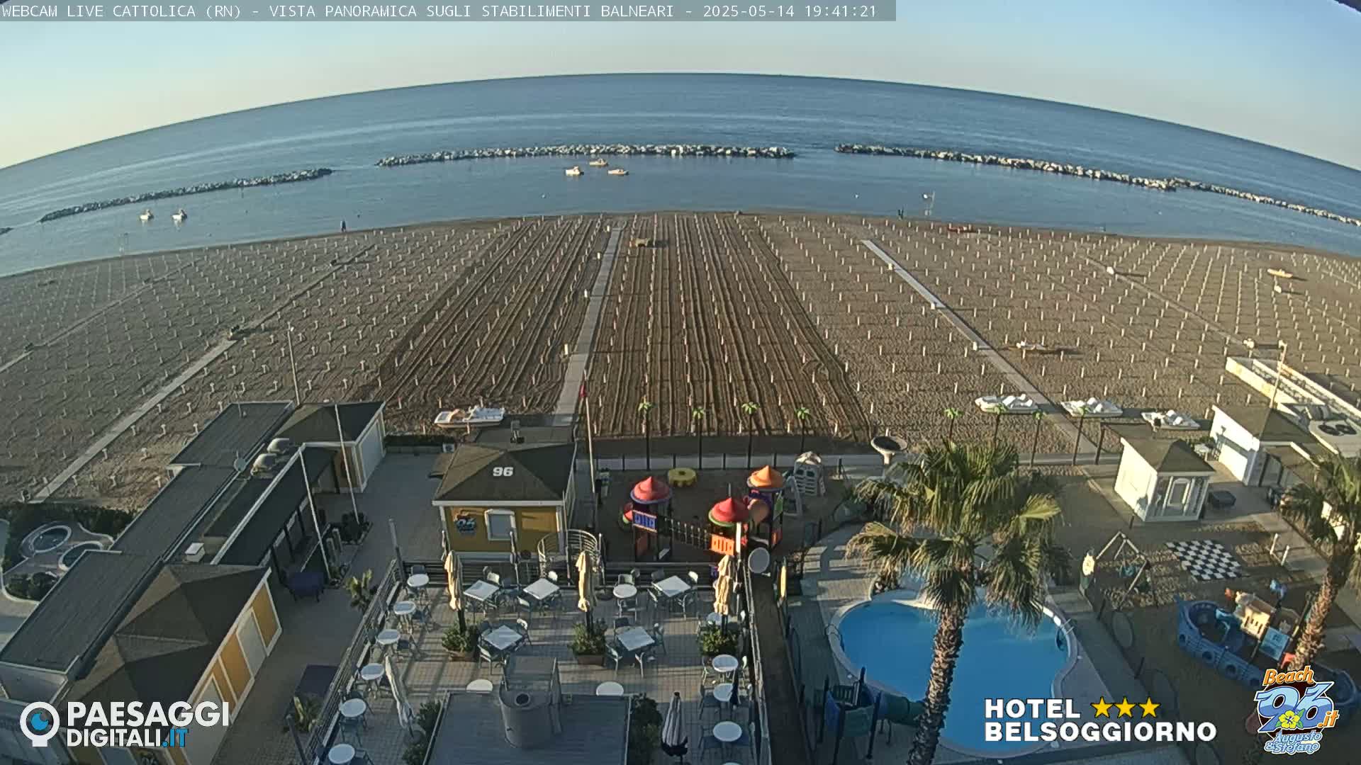 An aerial view of a beach with rows of beach umbrellas, a small playground, a swimming pool, and several buildings under a clear sky.