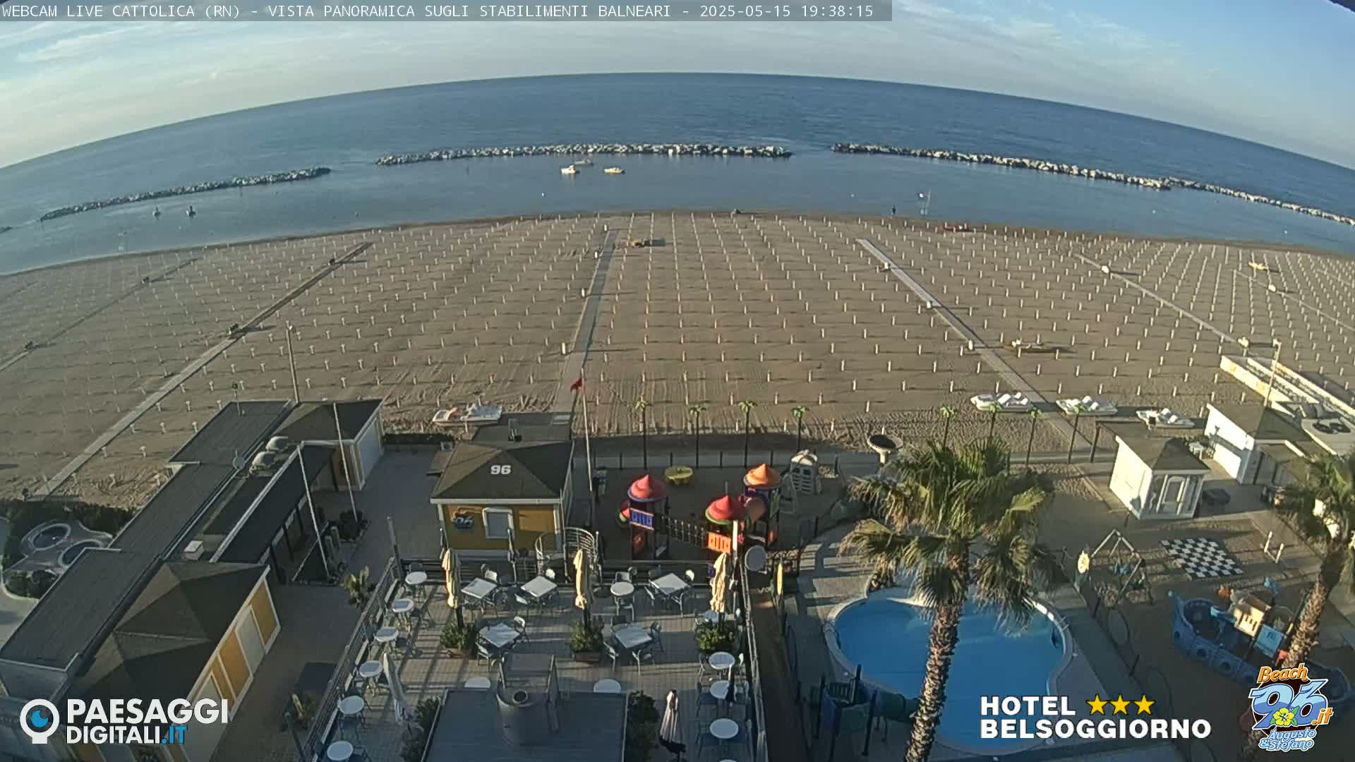 An aerial view shows a beach with rows of umbrellas and beach chairs, a small playground, a pool, and a cafe under a clear evening sky.