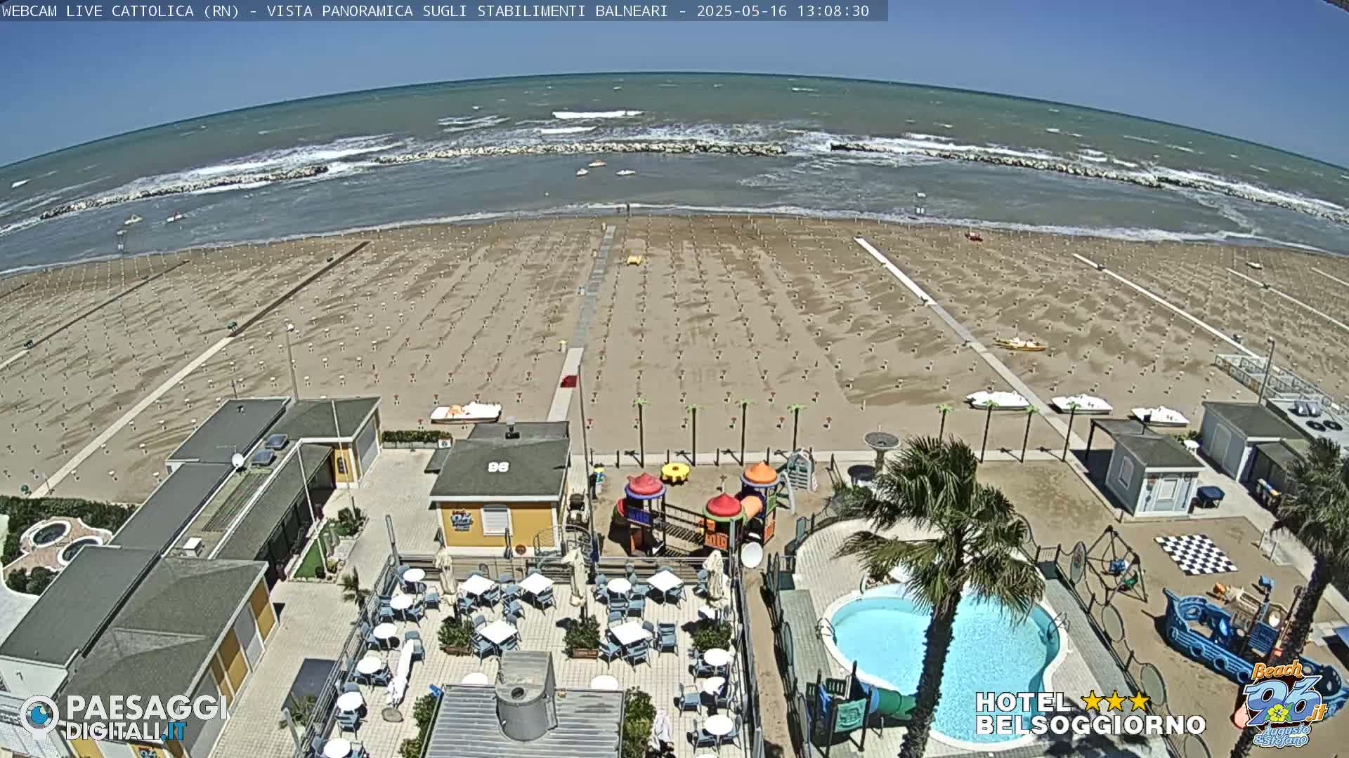 An expansive sandy beach with numerous umbrellas and beach equipment is visible, along with a beachside establishment featuring a pool, playground, and outdoor seating, all under a breezy sunny sky with a wavy ocean in the background.