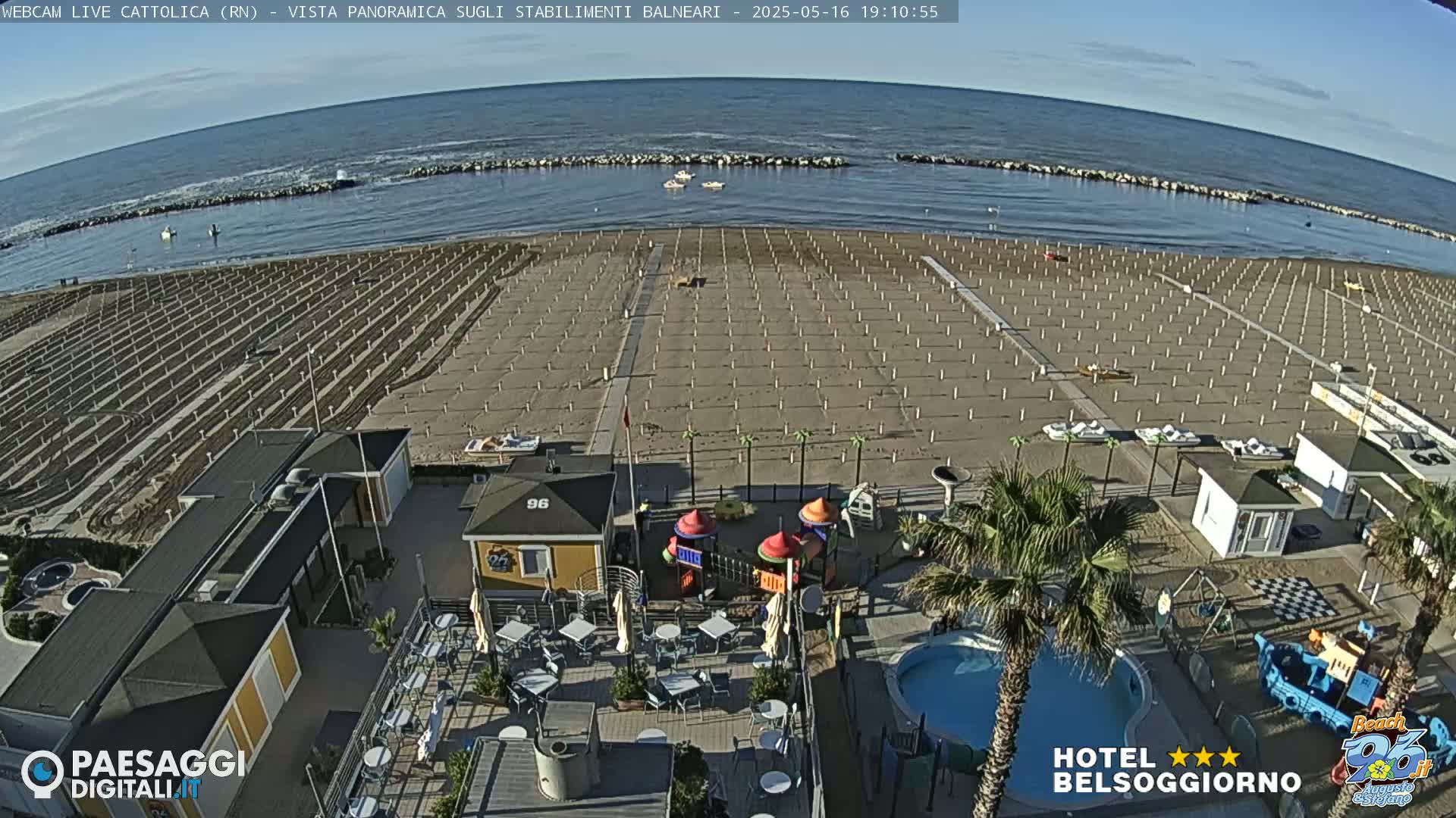An aerial view shows a beach with rows of beach umbrellas and chairs, a small playground, a pool, and a beachside restaurant under a clear sky.