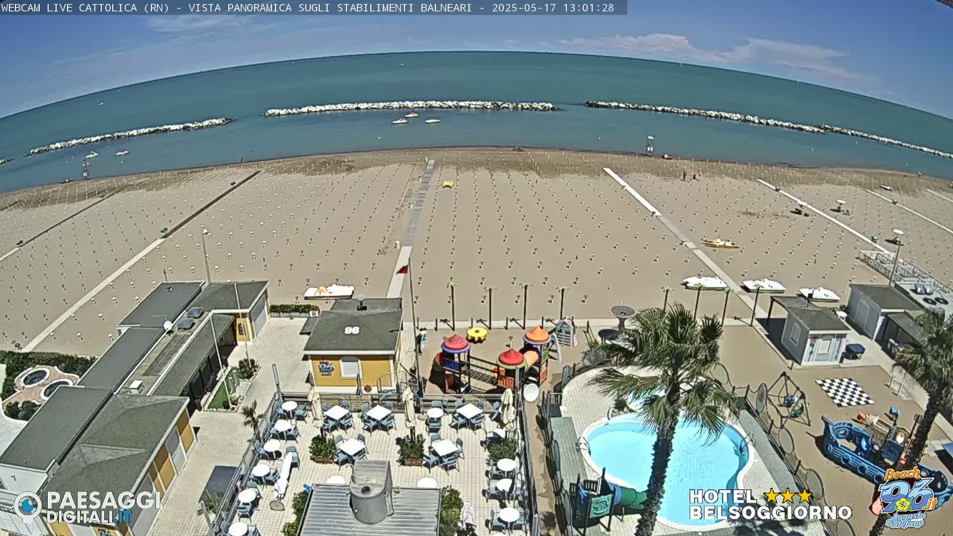 An aerial view of a beach with rows of empty beach umbrellas, a swimming pool, a playground, and a calm blue sea under a sunny sky.