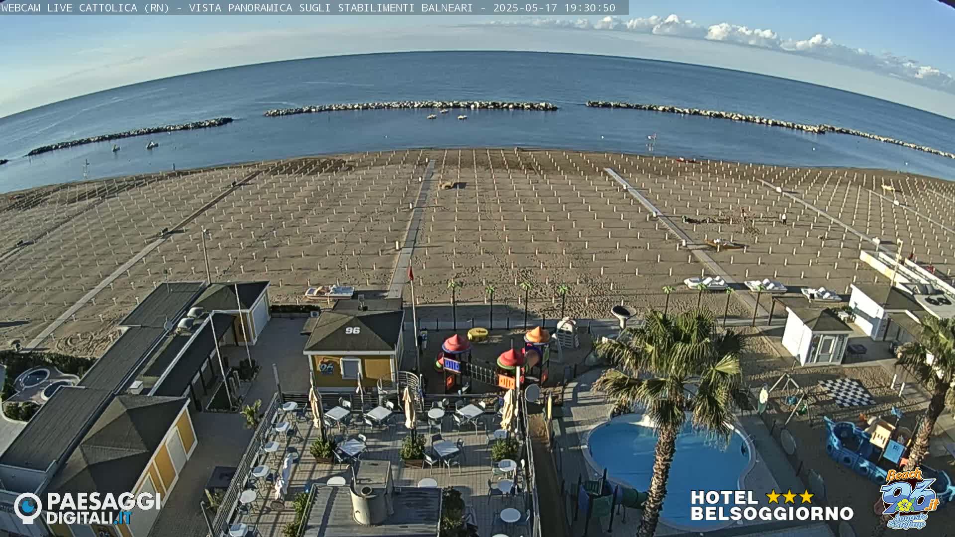An aerial view of a beach with rows of beach umbrellas and chairs, a small playground, a pool, and beachside buildings, on a mostly clear day.