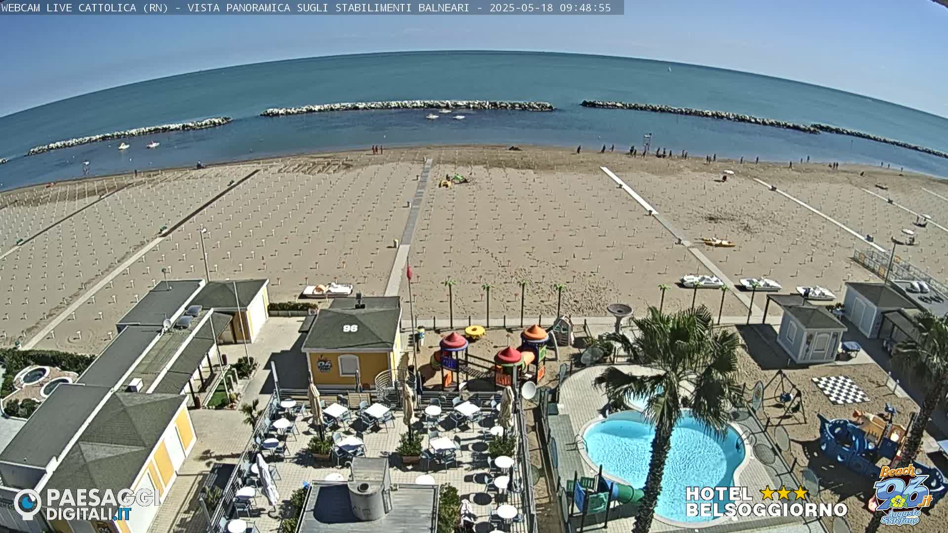 An aerial view of a beach with numerous umbrellas and beach chairs, a swimming pool, playground, and beachside restaurant, on a sunny day.