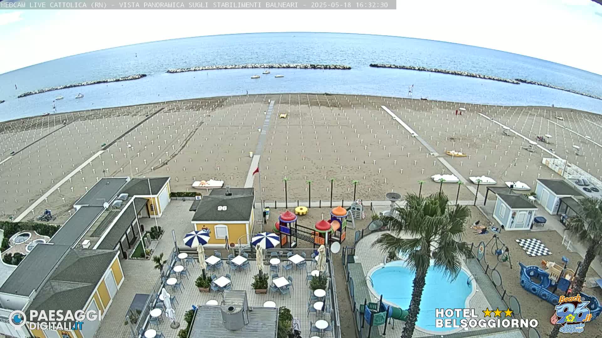 An overhead view shows a mostly empty sandy beach with rows of unoccupied beach umbrellas under a partly cloudy sky, next to a beachside hotel with a pool and playground.