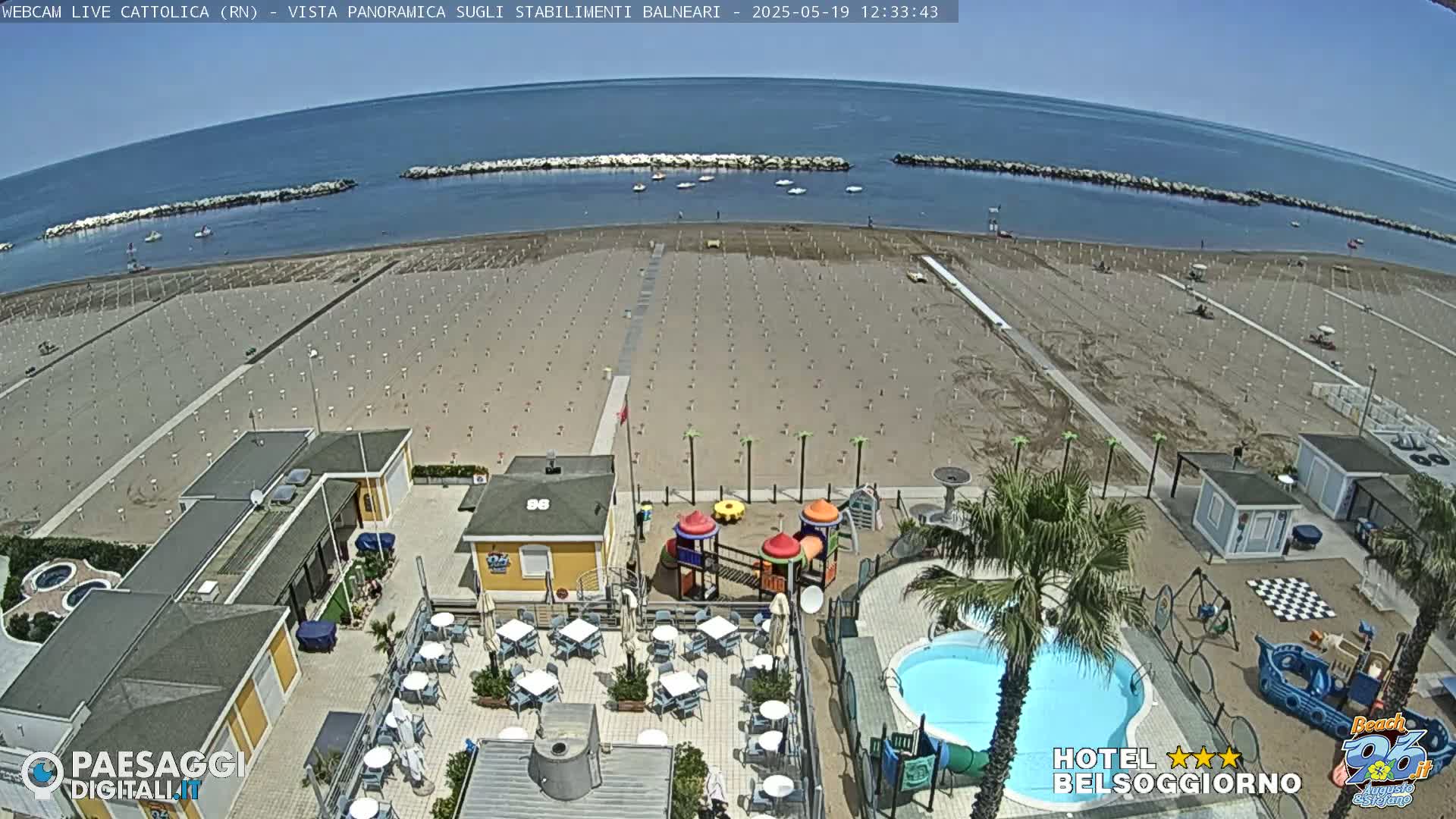 A beach with rows of empty umbrellas under a clear blue sky,  adjacent to a building with a small pool and playground.