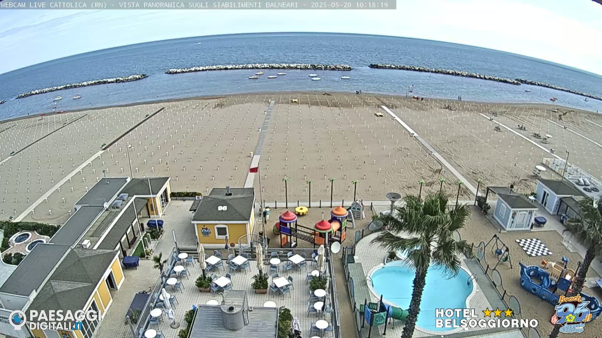 A mostly empty sandy beach with rows of closed umbrellas is seen from an elevated vantage point, overlooking beachside buildings, a small playground, a swimming pool, and calm blue water under a clear sky.