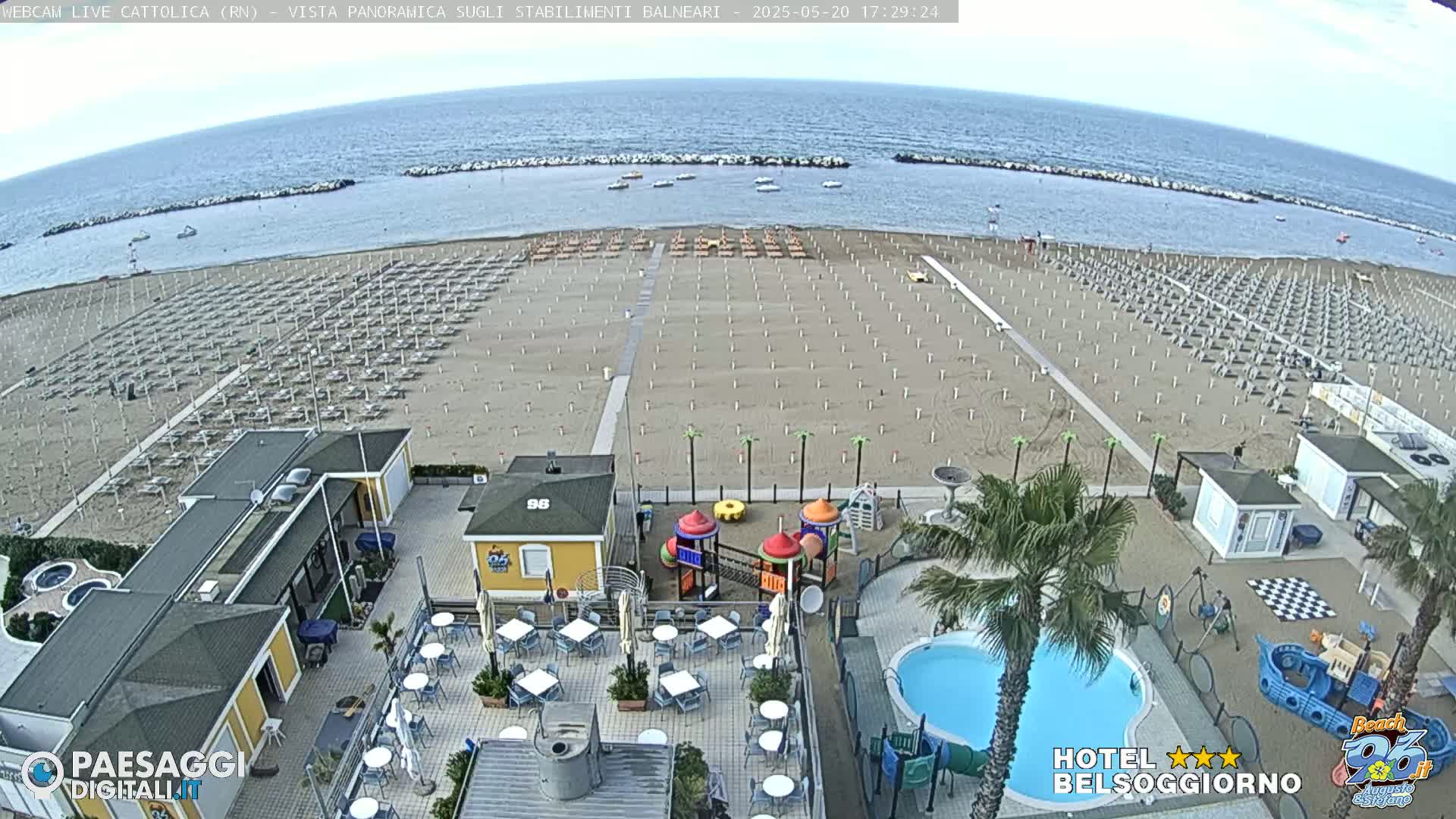 An aerial view of a sandy beach with numerous umbrellas and beach chairs, adjacent to beachside buildings, a playground, and a swimming pool, under a mostly clear sky.