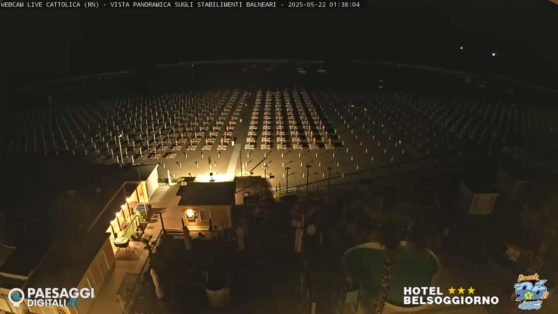 A nighttime aerial view shows a beach with rows of closed beach umbrellas and chairs, a building with lights on, and a small pool.