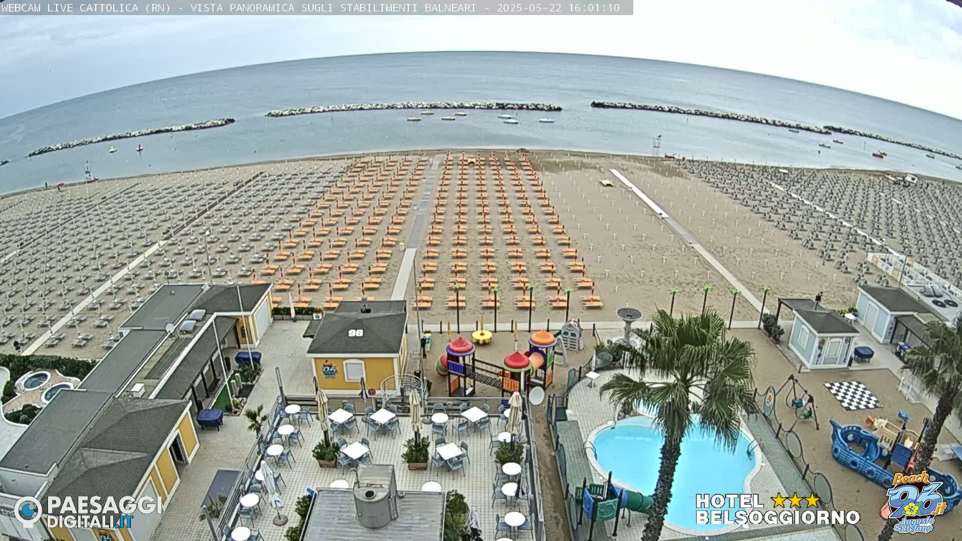 An overcast day at a beach with rows of empty beach umbrellas, a small playground, and a swimming pool next to a beachfront building.