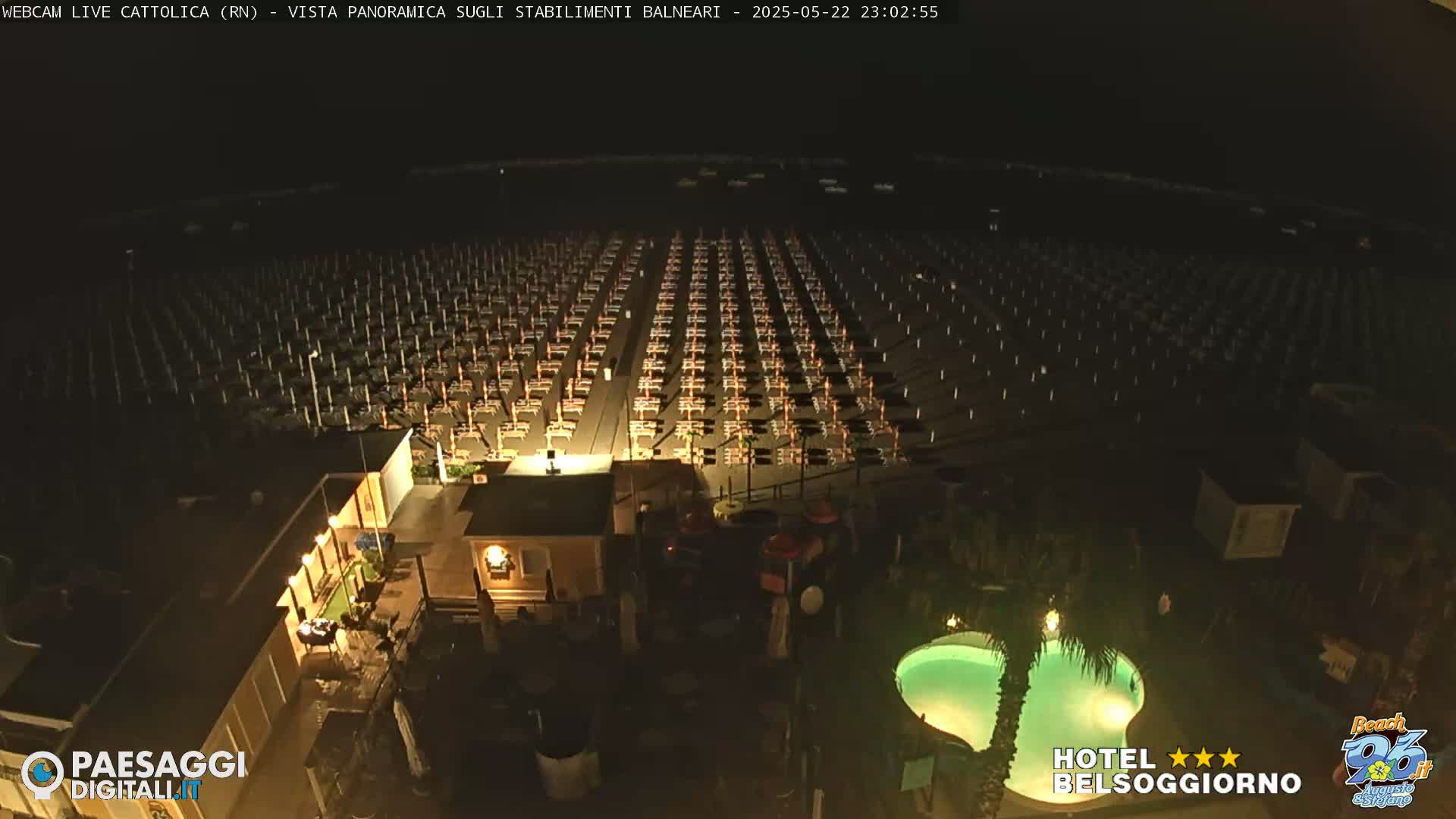 A nighttime aerial view of a beach shows rows of closed beach umbrellas and chairs, a small building, and a lit swimming pool.