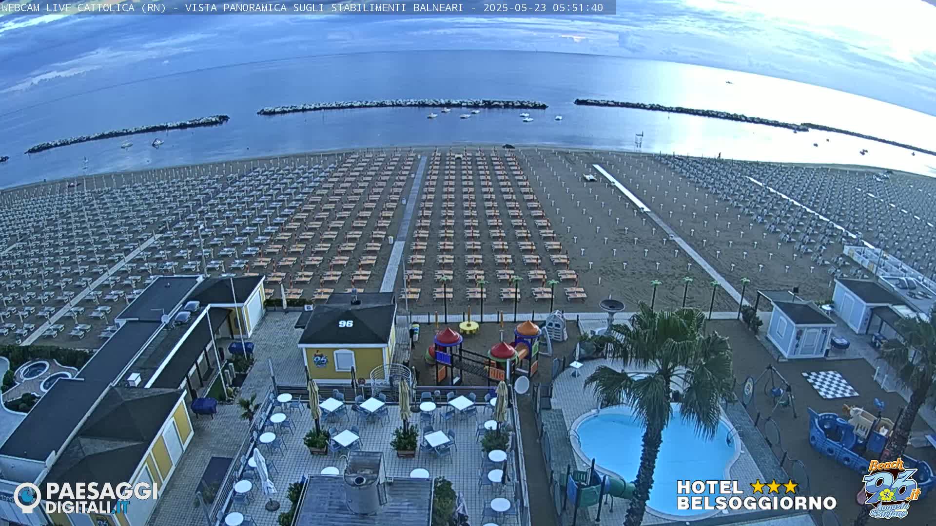 A beach with rows of empty beach umbrellas and lounge chairs is visible from an elevated viewpoint, overlooking a beachside establishment with a pool and playground, under a mostly clear sky.