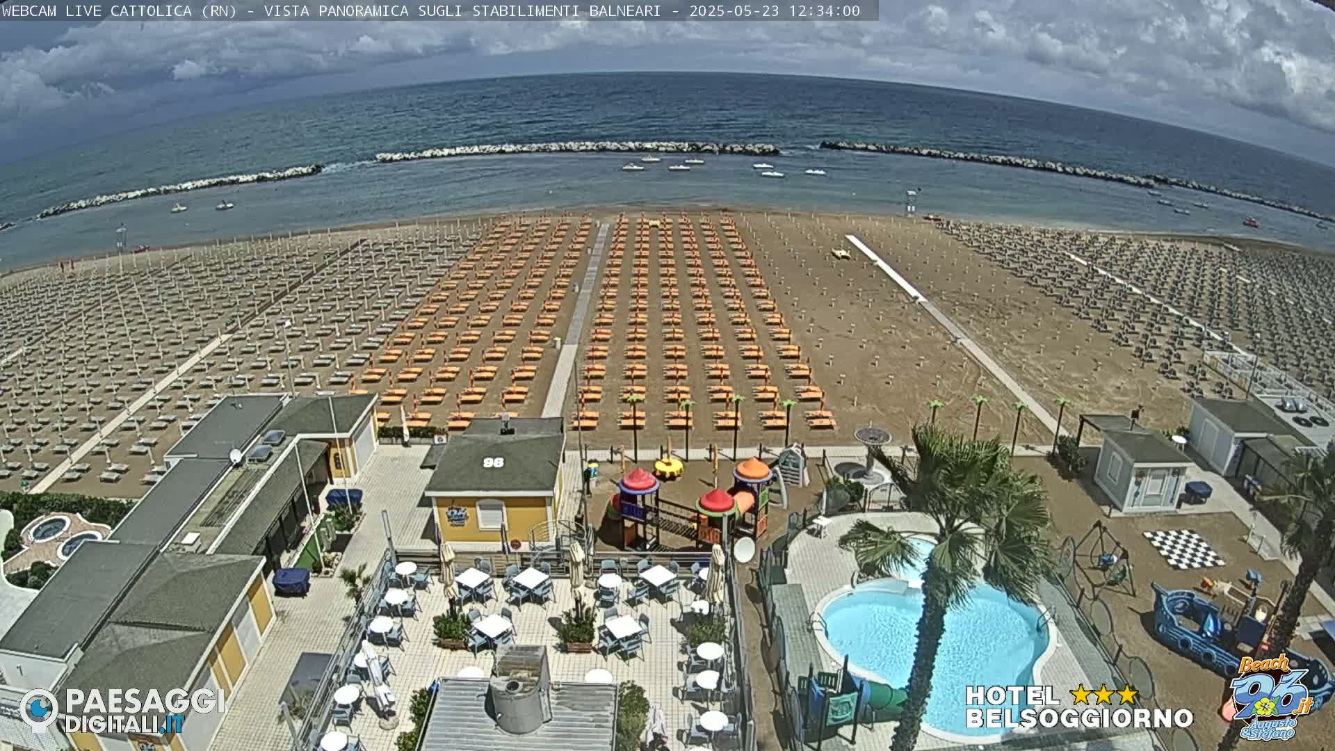 A beach with rows of orange beach umbrellas, a playground, a swimming pool, and beachside buildings is visible under a partly cloudy sky.