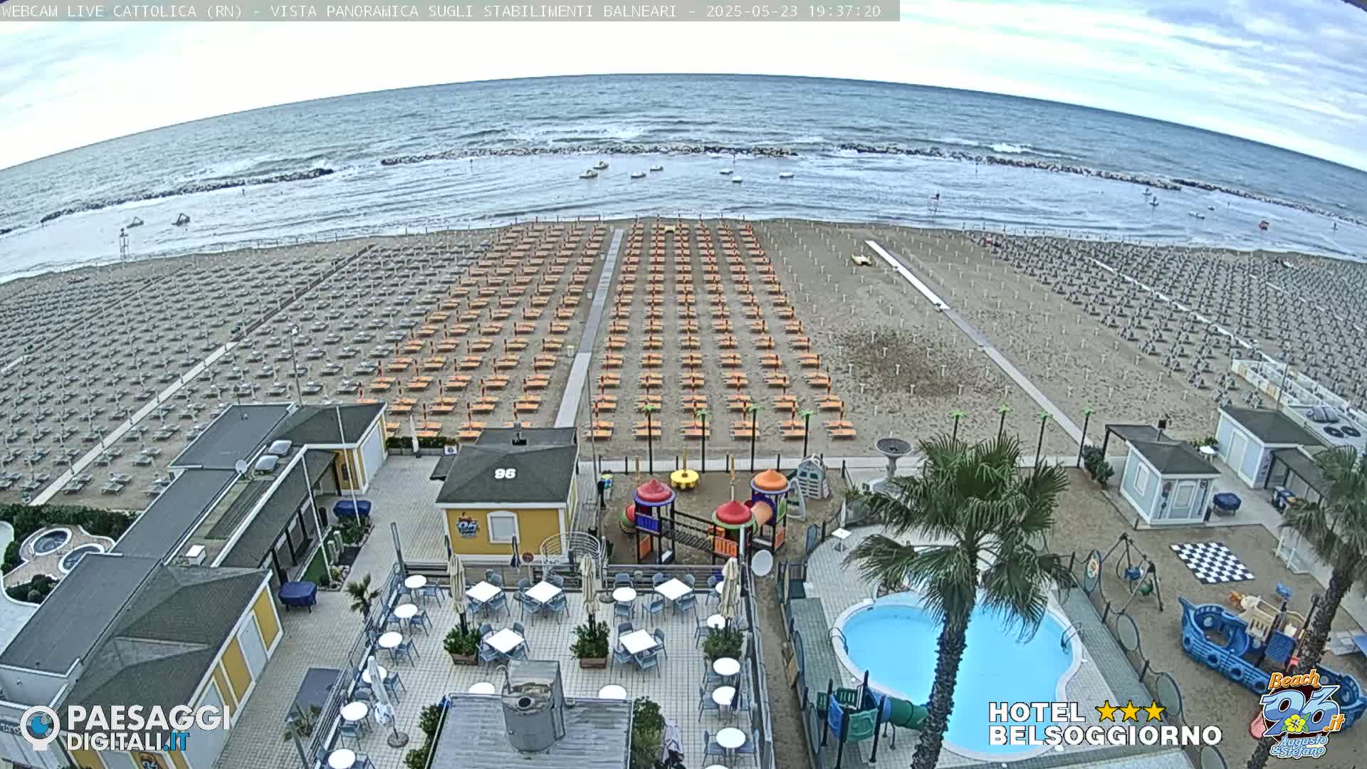 An overcast day at a beach shows rows of empty beach umbrellas and lounge chairs, a small playground, a swimming pool, and beachside buildings.