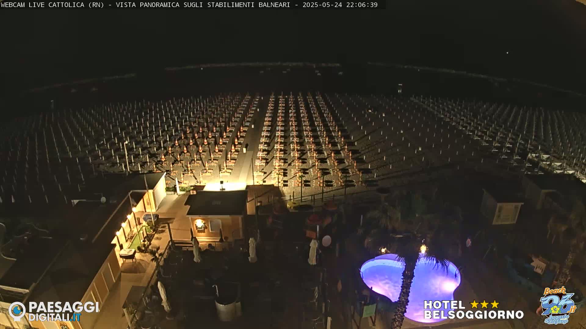 A nighttime, aerial view of a beach with rows of closed beach umbrellas and chairs, a small illuminated pool, and several buildings.