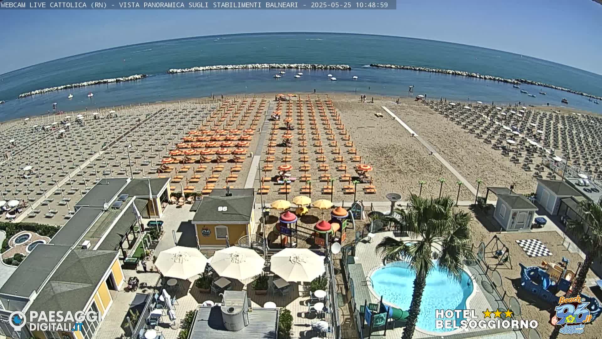 A beach with numerous orange umbrellas and lounge chairs, a swimming pool, playground equipment, and a calm sea under sunny skies is visible from an aerial perspective.