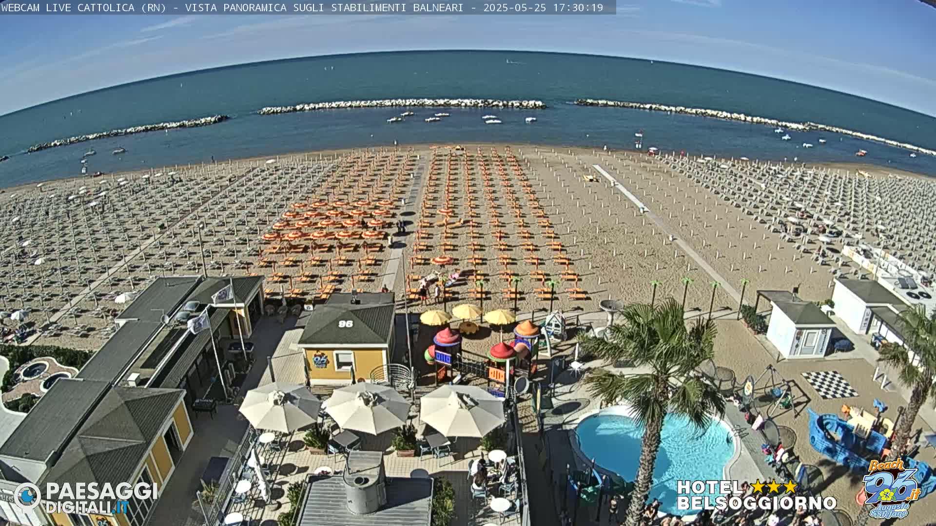 A sandy beach with rows of orange umbrellas and lounge chairs stretches along a calm blue sea under a sunny sky, and beachside amenities include a playground, a swimming pool, and beachside buildings.