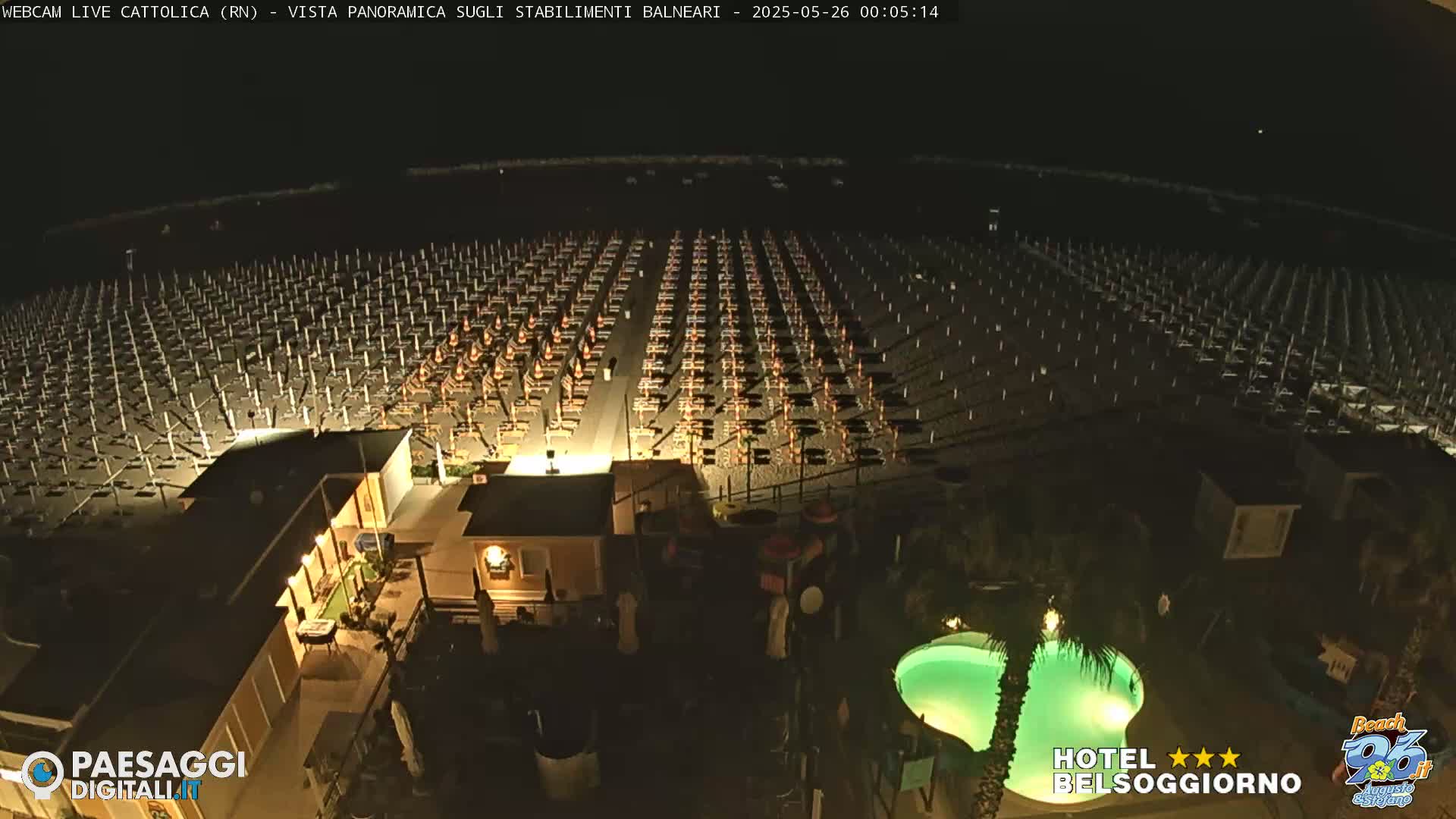 A nighttime, high-angle view of a beach at night showing numerous closed beach umbrellas and chairs, along with a small illuminated swimming pool and nearby buildings.