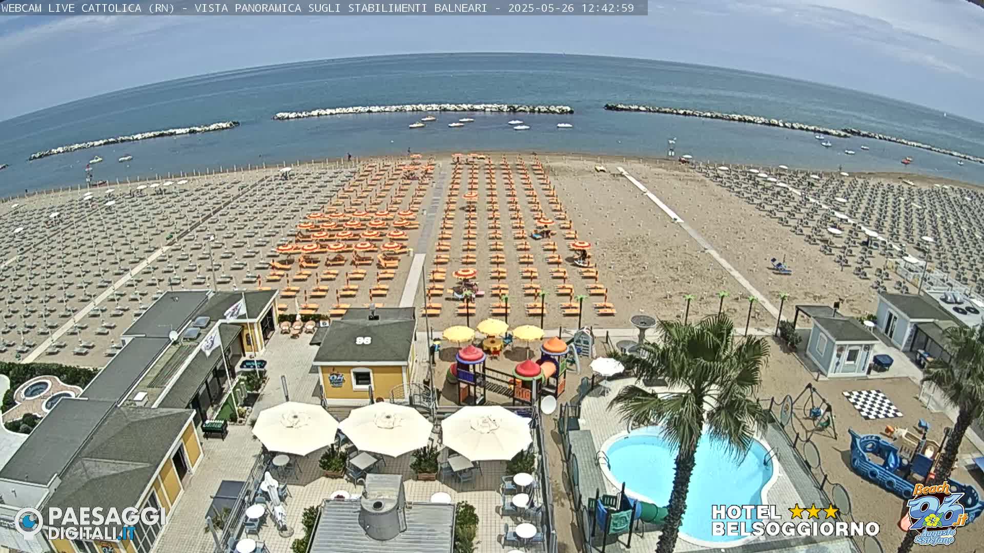 An aerial view shows a sandy beach lined with rows of beach umbrellas and lounge chairs, a beachfront area with a swimming pool and playground, and calm blue ocean beyond.