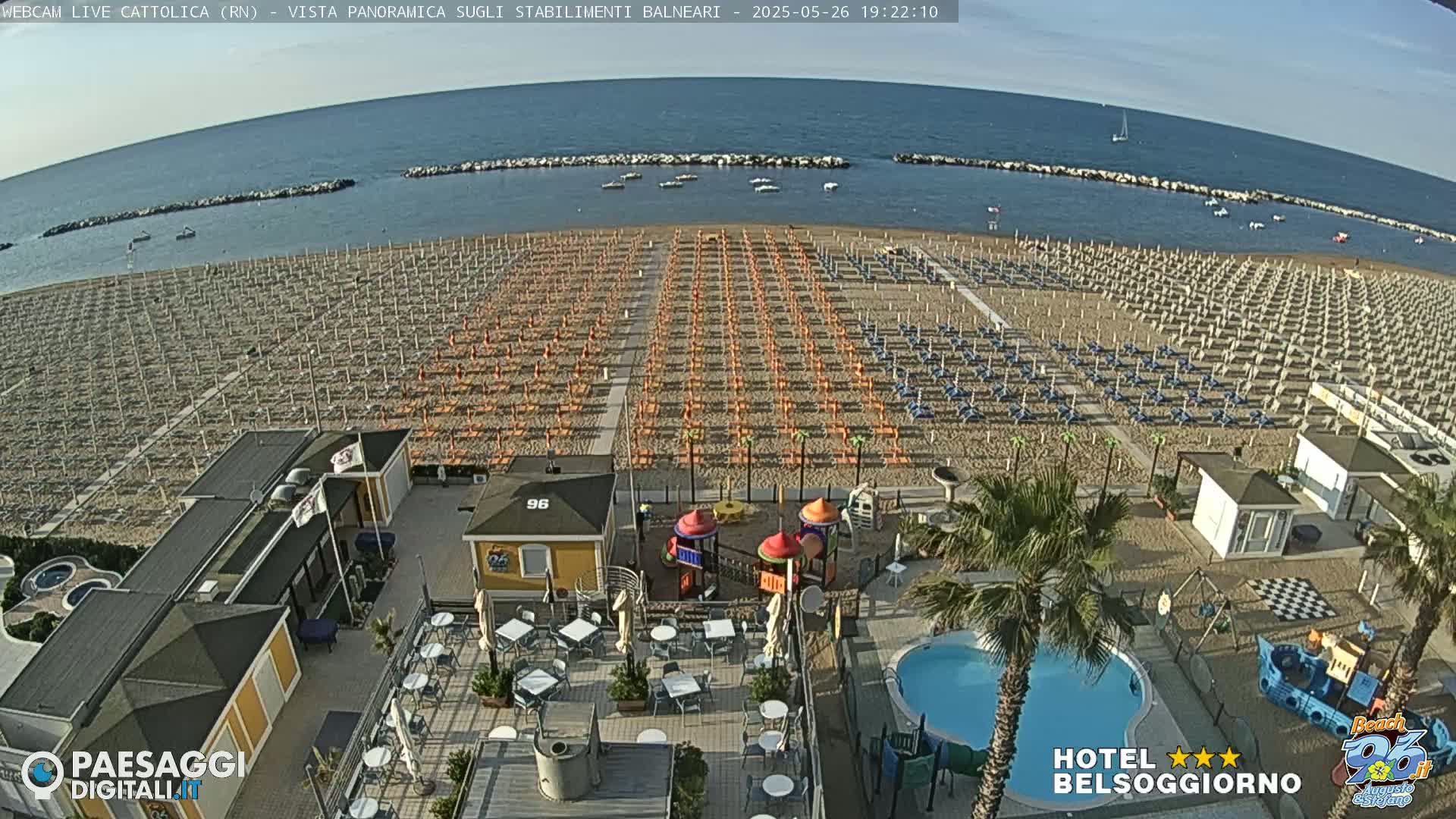 A beach with rows of umbrellas and beach chairs, a small playground, a swimming pool, and beachside buildings is visible under a clear sky.
