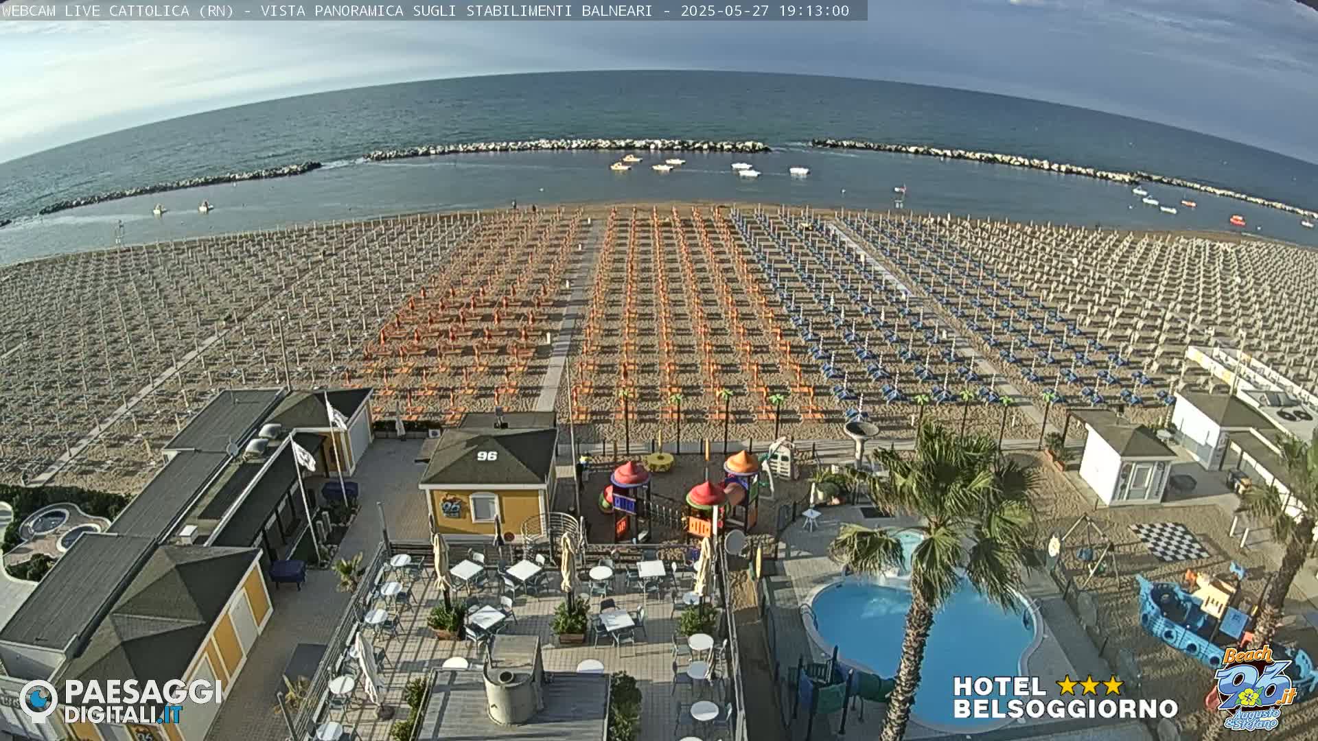 A beach with numerous rows of umbrellas and beach chairs, a small playground, a swimming pool, and some beachside buildings is visible under a clear, sunny sky.