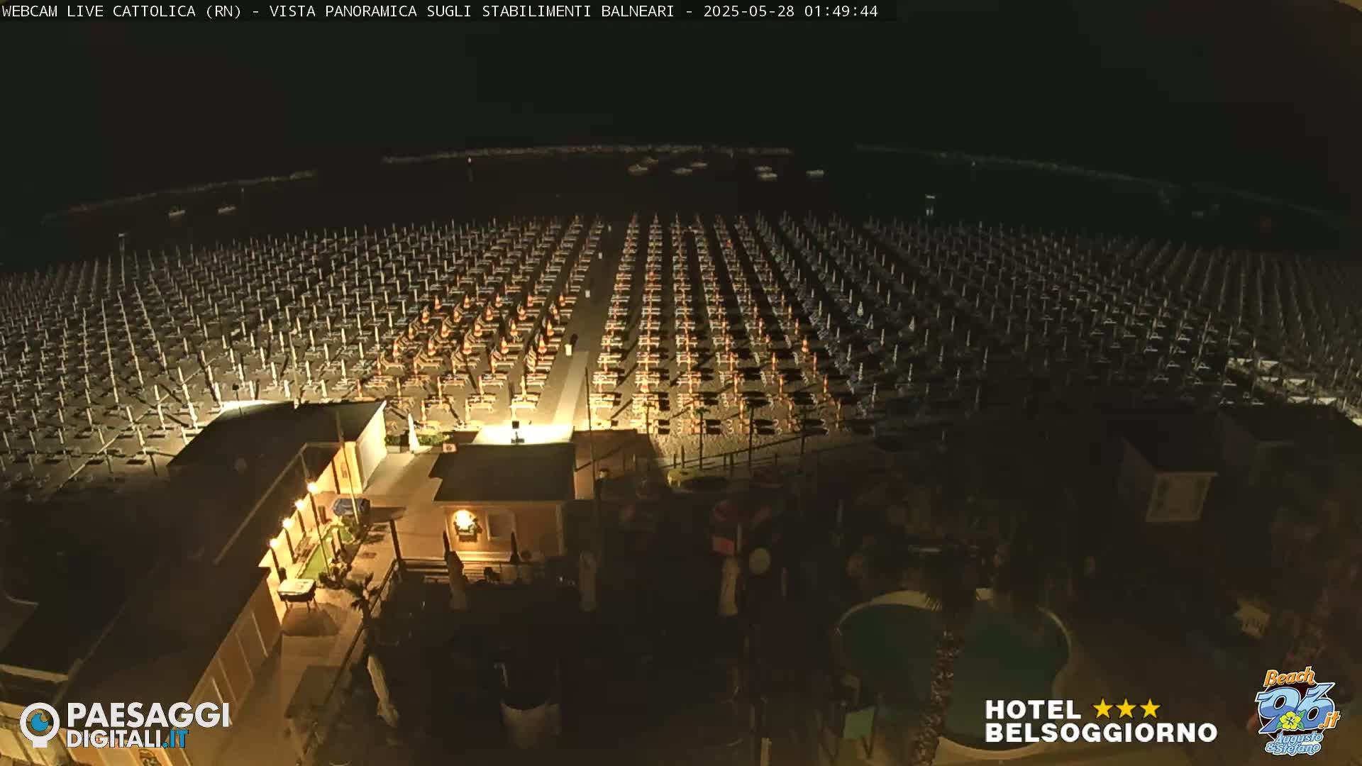 A nighttime, aerial view of a beach with rows of closed beach umbrellas and lounge chairs, and a small building with lights on.