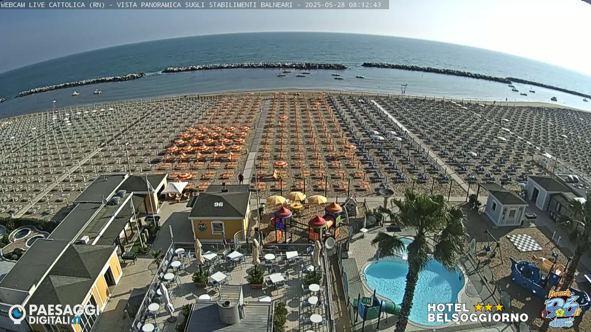 A beach with numerous rows of umbrellas and lounge chairs, a small pool, and a playground is visible under a sunny sky.