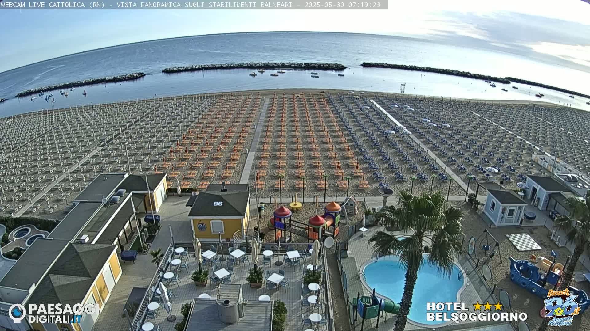 An aerial view shows a beach with numerous rows of beach umbrellas and lounge chairs, a swimming pool, playground equipment, and several buildings on a partly cloudy day.
