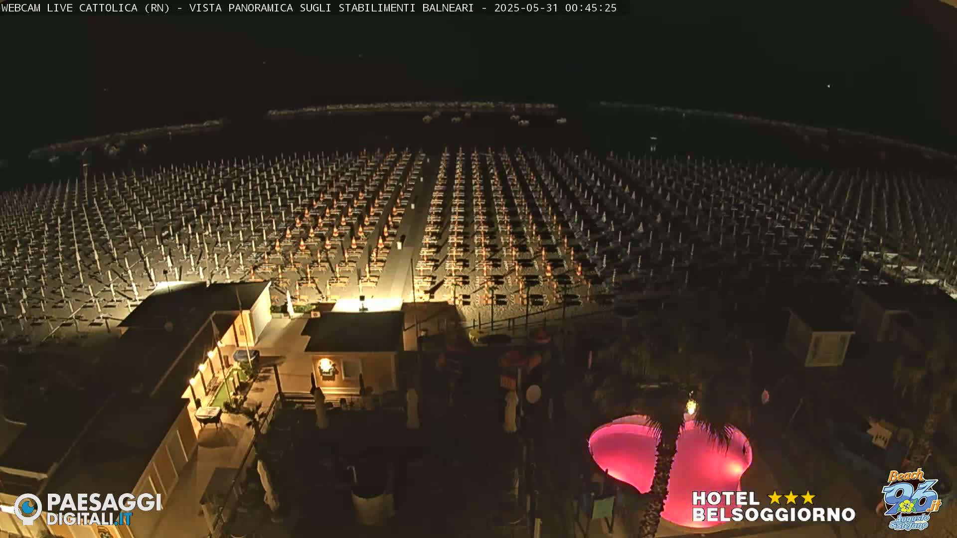 A nighttime aerial view of a beach at night, showing numerous closed beach umbrellas and chairs, and a pink-lit pool near a building.