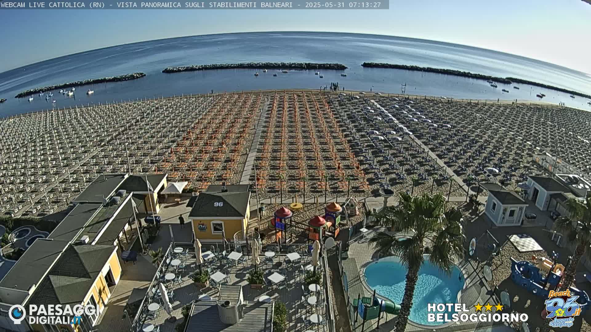 An aerial view shows a beach with rows of umbrellas and beach chairs, a small playground, a swimming pool, and several small buildings under a clear, sunny sky.