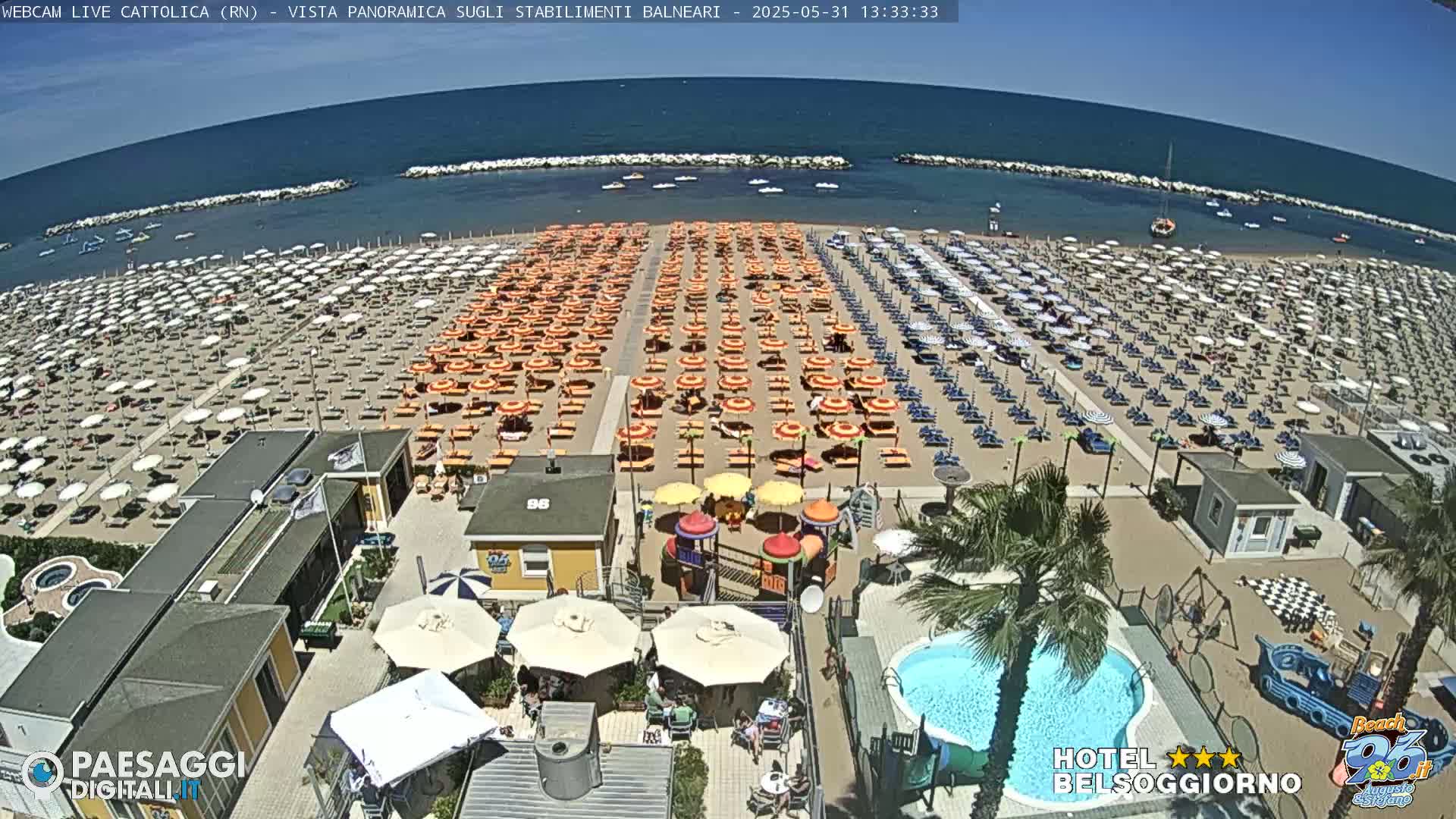 A sunny beach scene shows numerous umbrellas and lounge chairs on a sandy beach next to a calm ocean; a swimming pool and playground are visible closer to the camera.