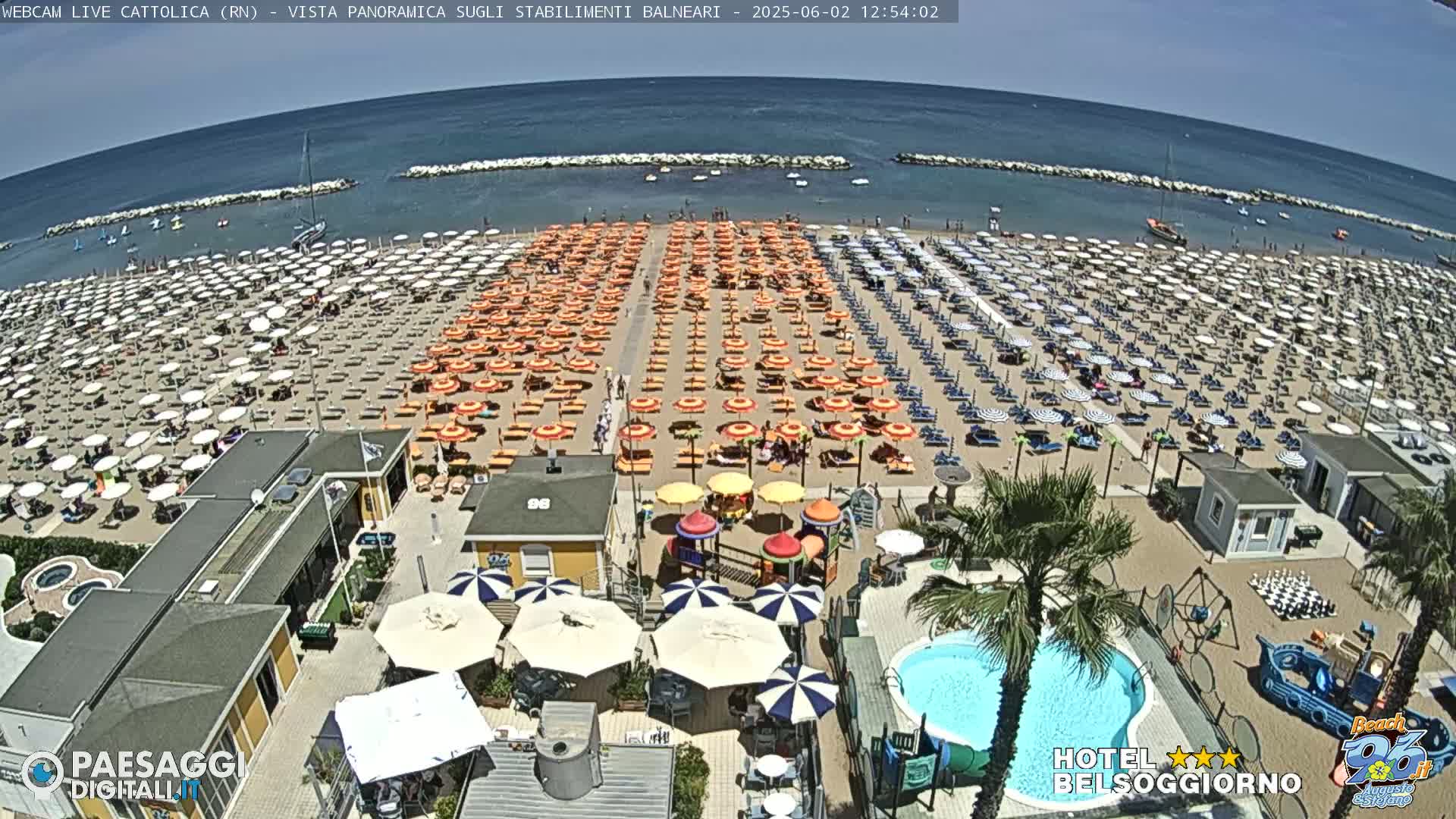 A beach with numerous orange and white umbrellas, beach chairs, a swimming pool, and a playground is shown under sunny skies.
