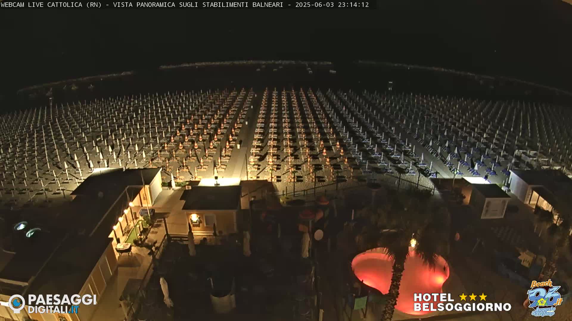 A nighttime, aerial view of a beach with rows of closed umbrellas and beach chairs, illuminated buildings, and a red-lit pool.