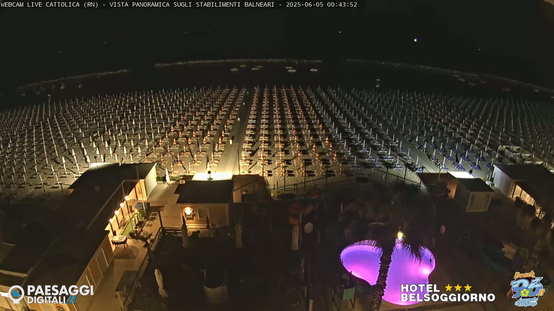 A nighttime aerial view of a beach with numerous closed umbrellas and lounge chairs, illuminated buildings, and a purplish-lit swimming pool.