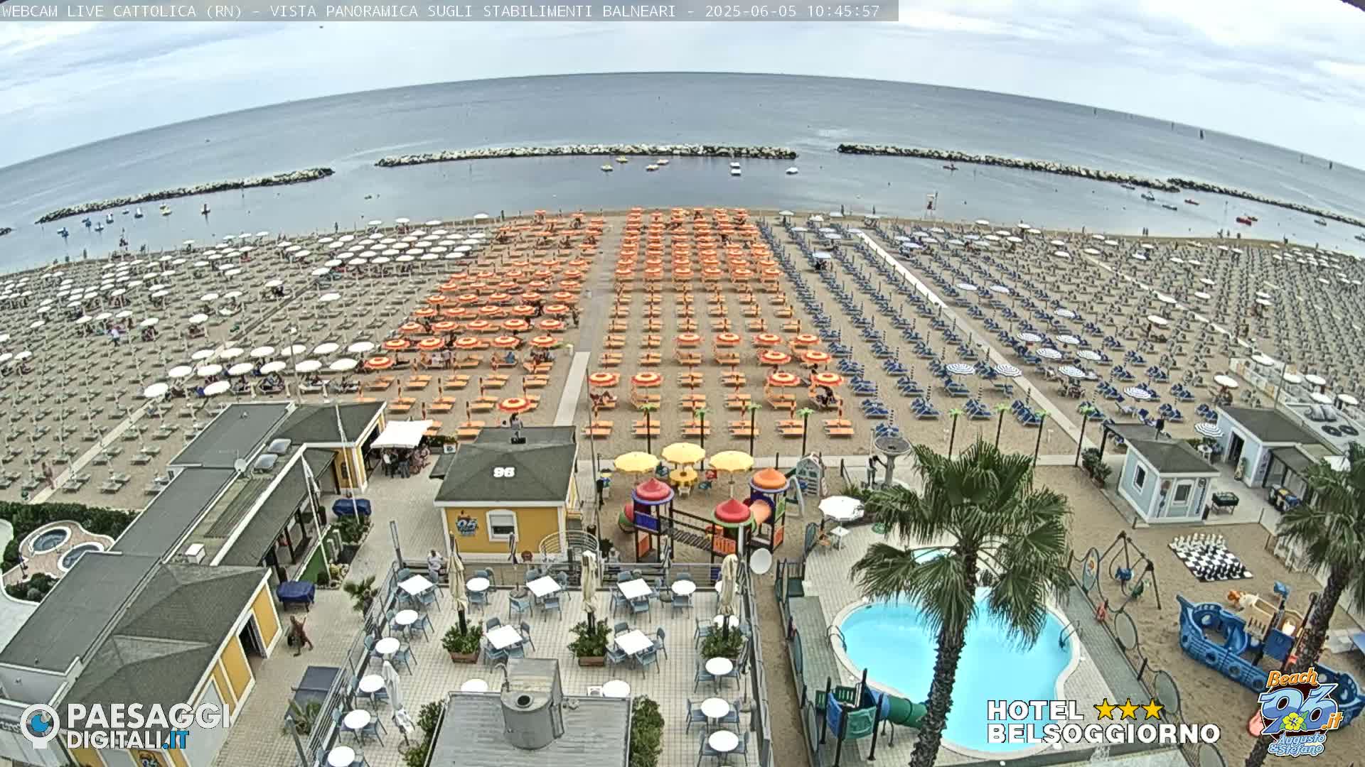 A beach with numerous rows of umbrellas and lounge chairs, a small playground, and a swimming pool is visible on a partly sunny day.