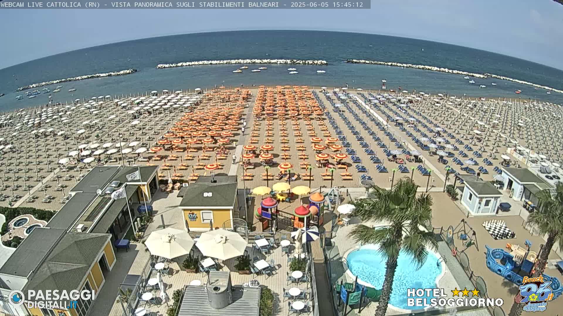 A sunny beach scene shows numerous beach umbrellas and lounge chairs arranged in rows, along with a small playground, swimming pool, and beachside buildings.