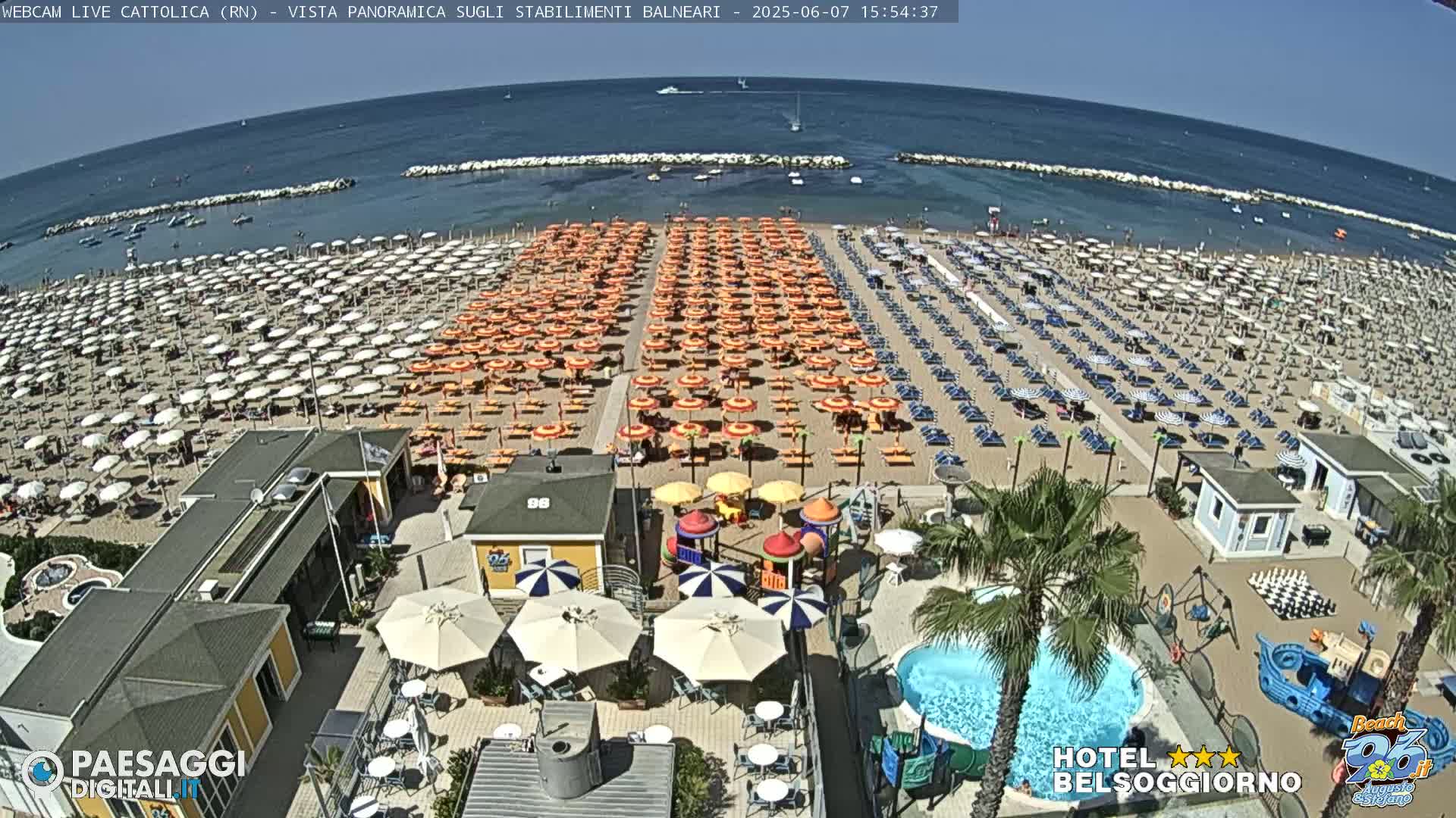 A sunny day at a crowded beach with numerous umbrellas and beach chairs, alongside a playground and swimming pool.