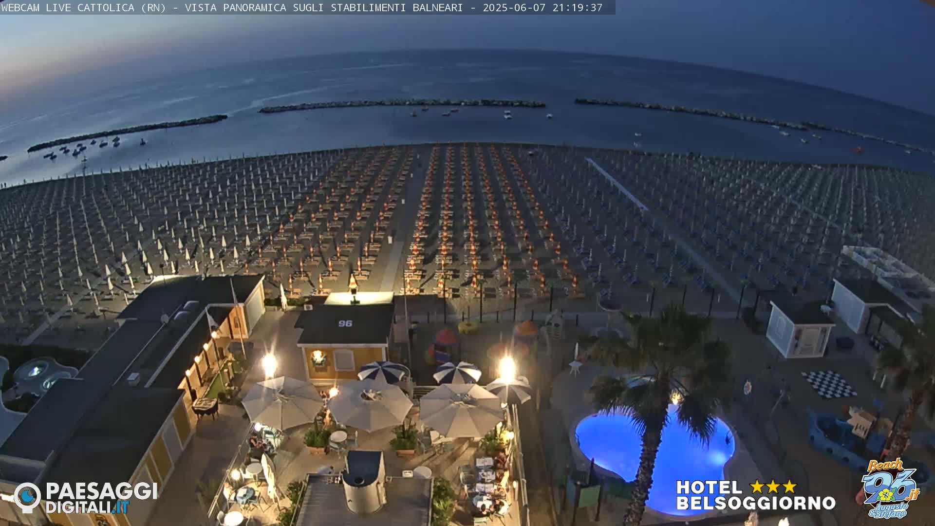 A beach at night, filled with rows of closed beach umbrellas, is viewed from above, showing a hotel pool and outdoor seating area in the foreground.