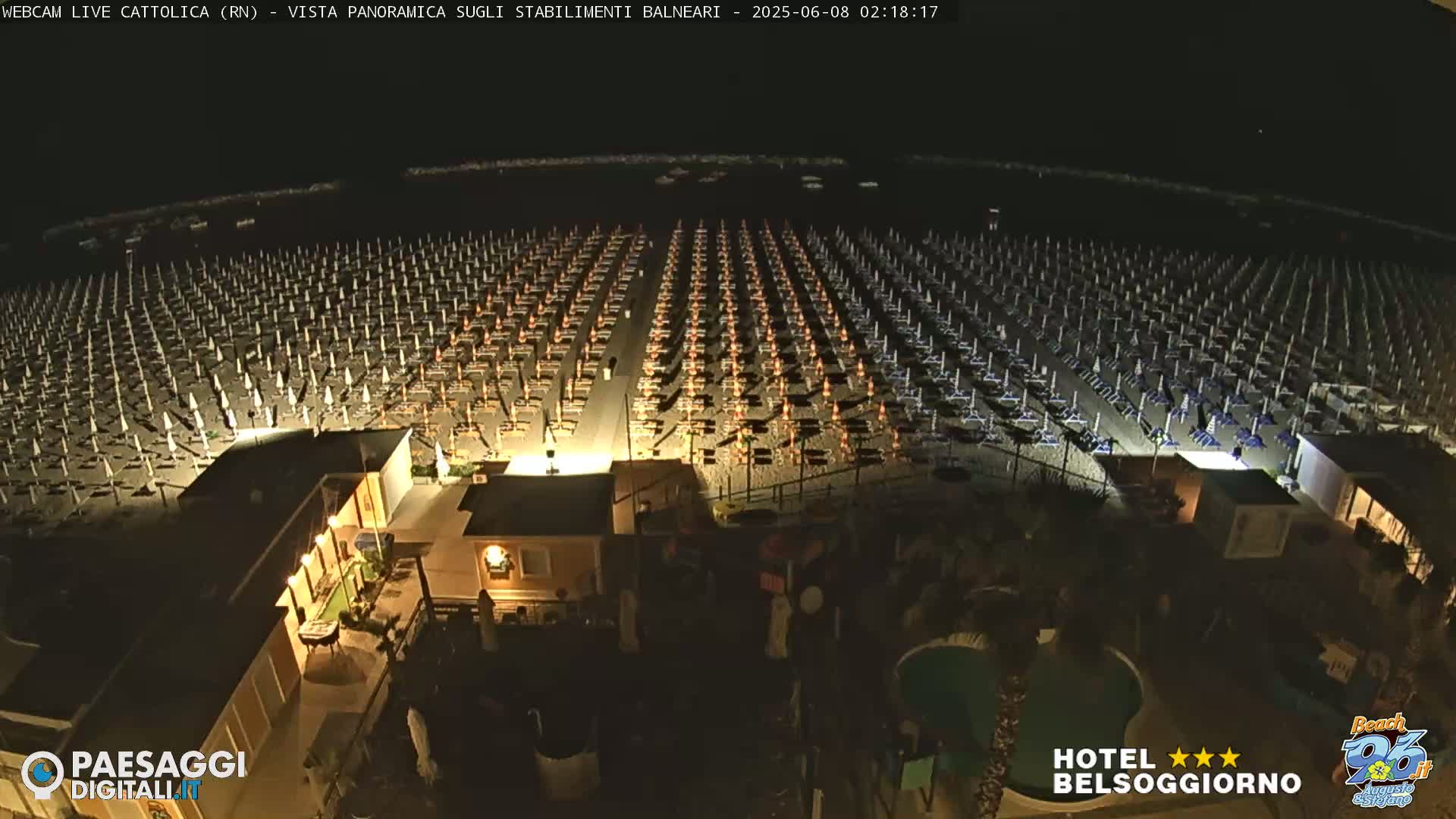 A nighttime, aerial view of a beach with hundreds of closed umbrellas and beach chairs illuminated, along with a small building and a swimming pool.