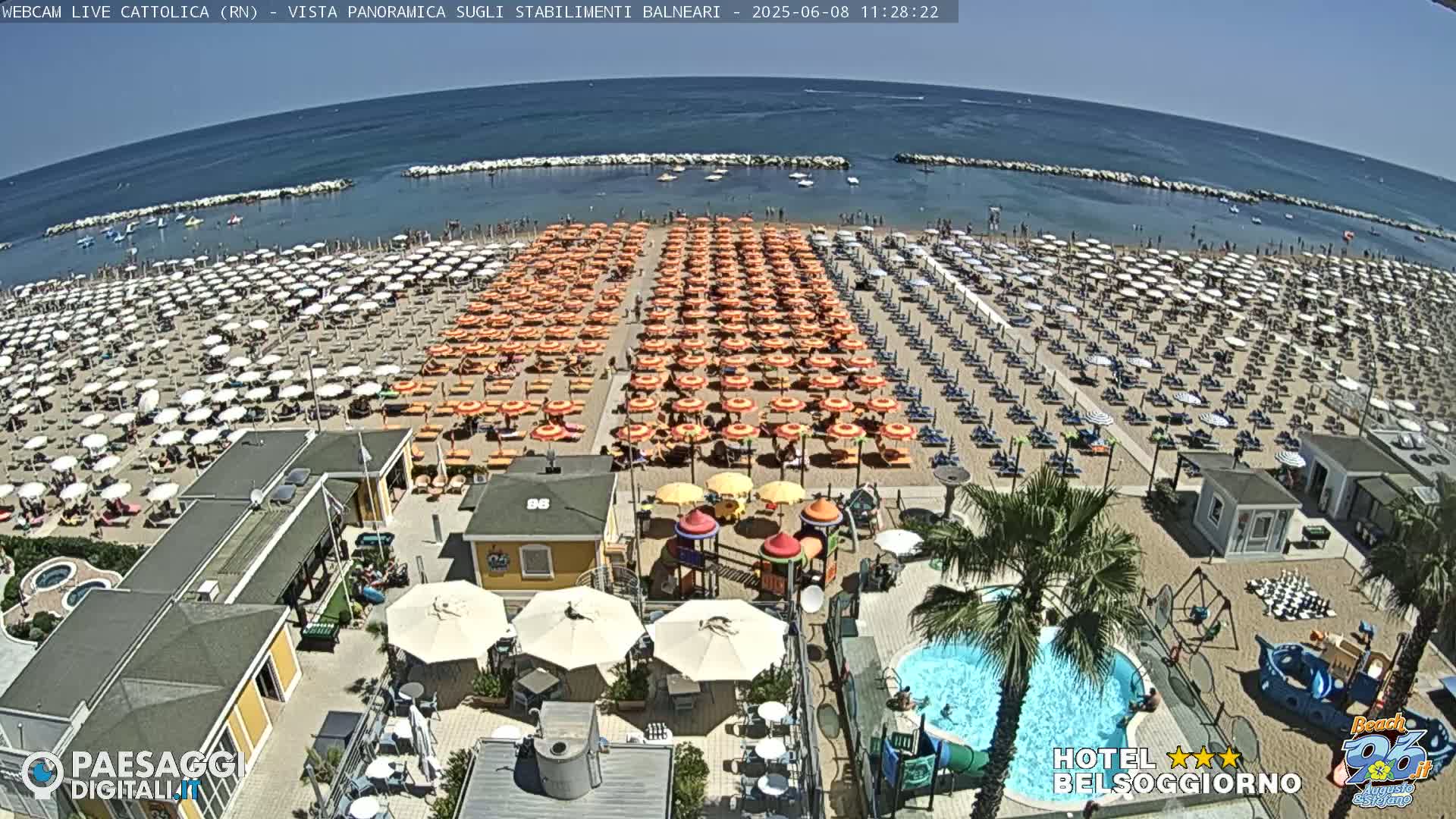 A sunny beach with numerous umbrellas and lounge chairs, a swimming pool, and a playground is shown, overlooking a calm ocean.